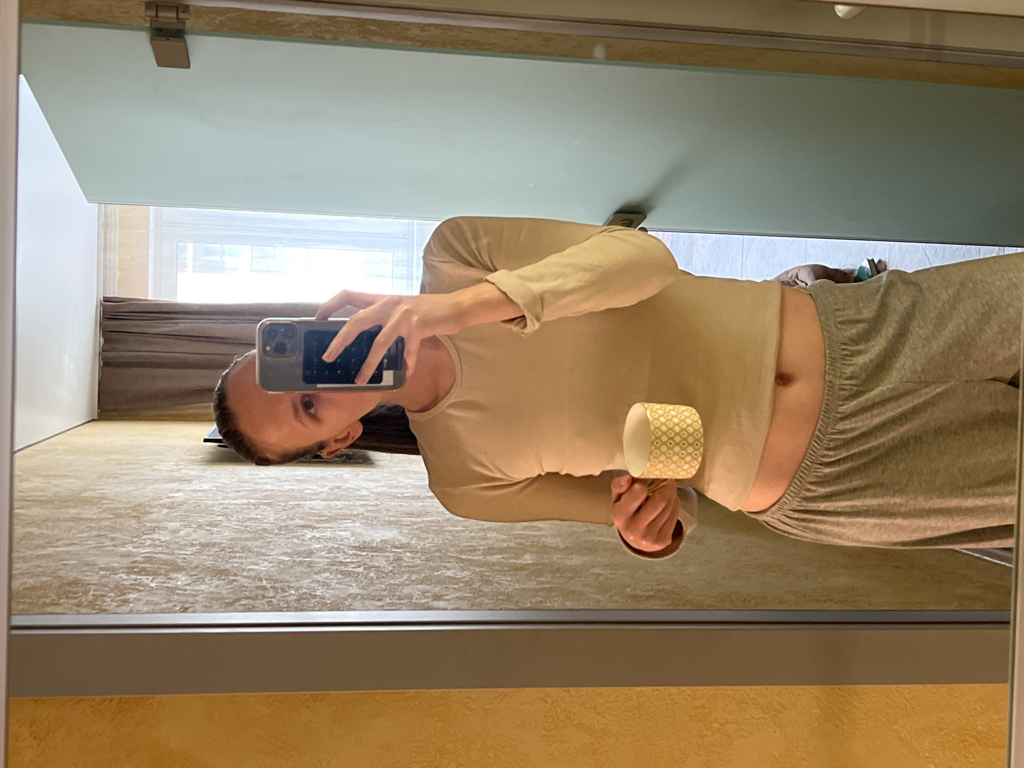 A woman is reflected in a mirror, taking a selfie with a phone. She wears a light tan long-sleeved shirt and gray sweatpants. She's holding a patterned cup in her other hand. The location appears to be indoors, with a window showing a view of what may be a city or residential area. There are brown curtains visible through the window. The floor has a patterned beige and brown design. Based on available information, I cannot definitively pinpoint the location as the city of Khimki, Russia.