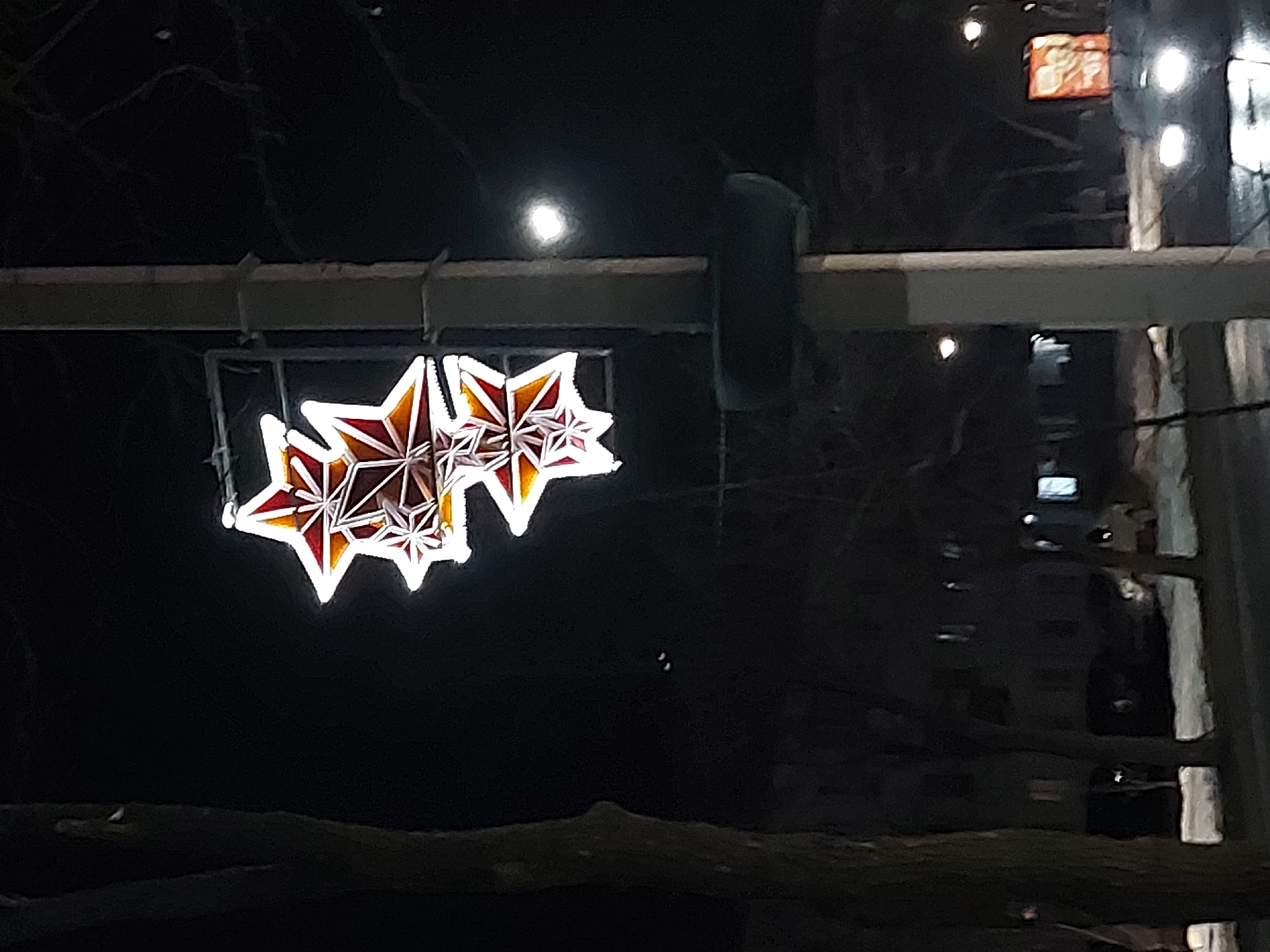 The image shows a night scene in what appears to be the city of Chaikovsky, Russia. Three star-shaped decorations, illuminated with a combination of white and reddish-orange light, are suspended from a horizontal beam above. The background is dark, with a blurred cityscape visible, including buildings with lights in some windows. A street or sidewalk is visible in the foreground. The presence of the star decorations suggests a holiday or celebratory occasion.
