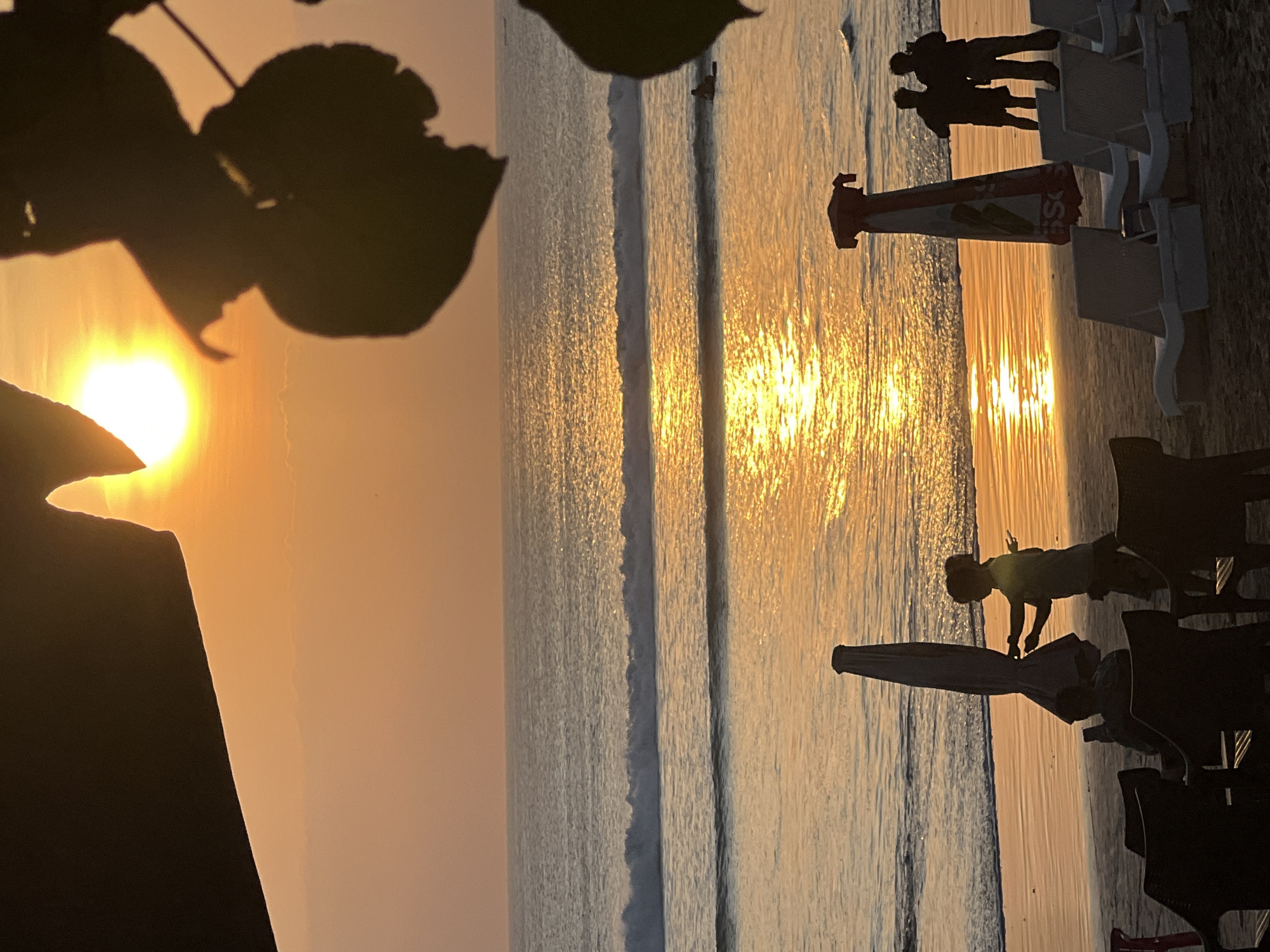 A silhouette of a sunset on Kuta Beach, Indonesia, shows the sun as a bright orb just above the horizon, casting a golden reflection on the ocean. Waves gently roll towards the shore. On the beach, several people are visible. In the foreground, a young child stands near a folded beach umbrella. Further back, two adult figures stand together. In the middle ground, a person is lying on a surfboard in the water. Beach chairs are scattered on the sand, some occupied by silhouetted figures. The scene is framed by dark foliage in the upper right corner and a dark, curved shape in the upper left corner. The overall atmosphere is peaceful and serene, typical of a Kuta Beach sunset.