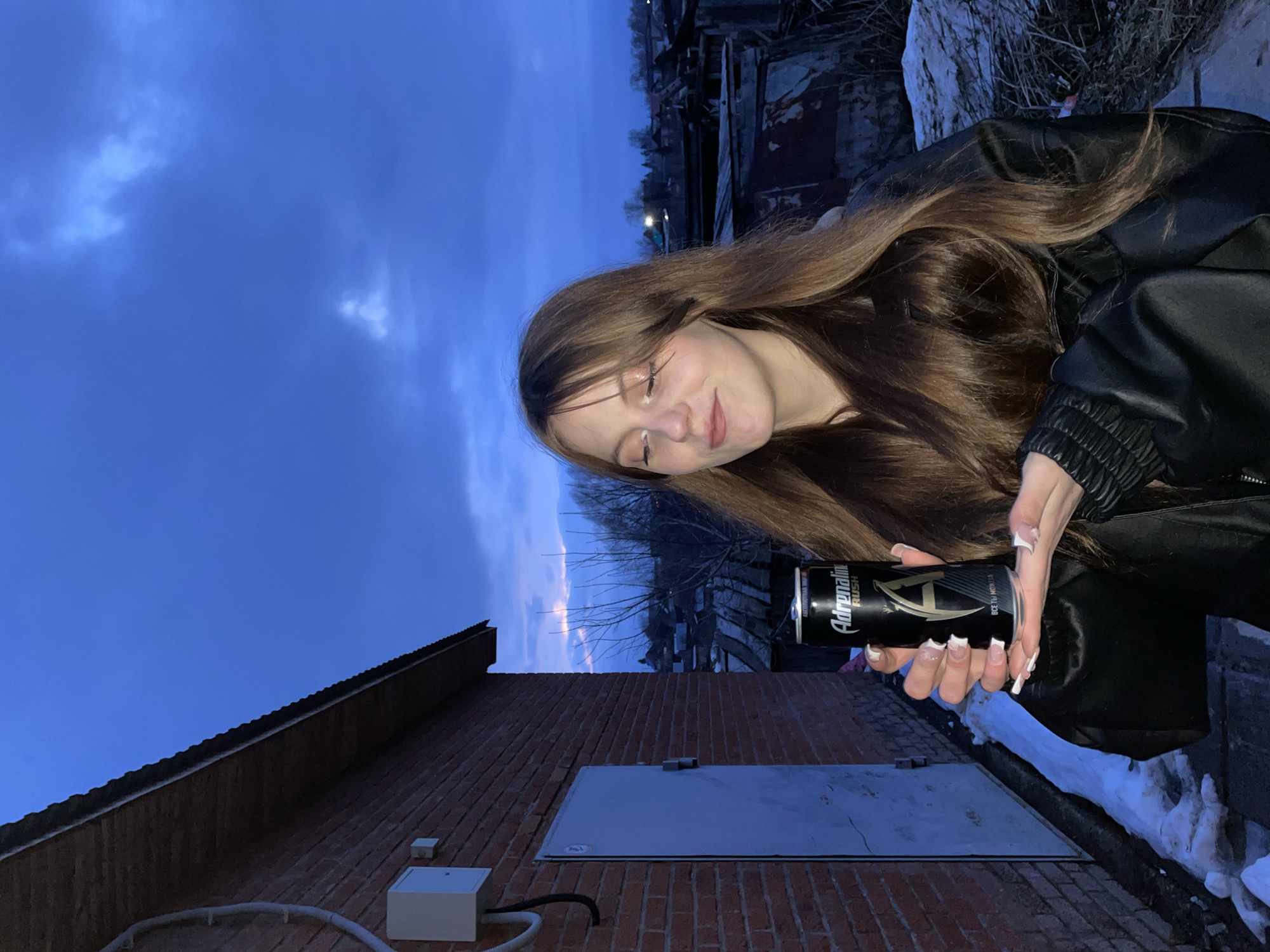 A young woman with long, brown hair is holding a can of Adrenaline Rush energy drink. She is wearing a black leather jacket. The background shows a brick building, a metal door, snow, and a cloudy sky. The location is Фоки, Russia.
