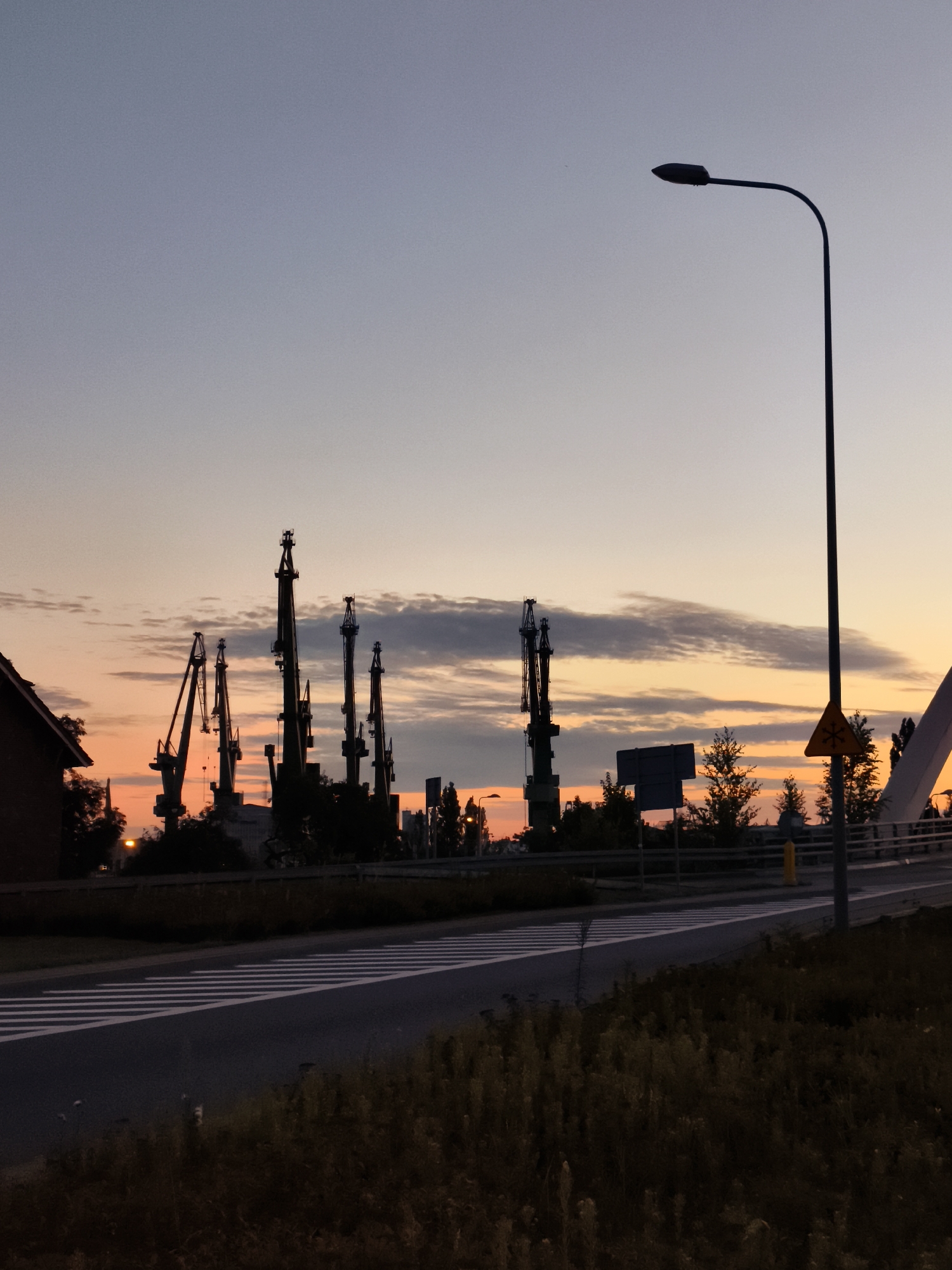 The image depicts a dusky industrial waterfront scene in Gdańsk, Poland, likely during sunrise or sunset. Dominating the background are several large, silhouetted dock cranes, their skeletal structures stark against a sky painted with hues of orange, pink, and pale blue. Wisps of clouds are visible, catching the fading or emerging light.
In the foreground, a dark asphalt road cuts across the frame, marked with a prominent white pedestrian crossing. To the right of the road, a modern street lamp with a curved arm stands tall, its dark silhouette a sharp contrast to the sky. Further to the right, a white architectural element, possibly part of a bridge or a structure, curves upwards. A yellow triangular warning sign with a black snowflake symbol is also visible on this side.
On the left side of the image, the corner of a dark-colored building, possibly made of brick, is partially visible. Between the building and the cranes, trees and other vegetation create dark masses. The overall atmosphere is quiet and still, typical of the early morning or late evening in a port city. The lighting suggests a low sun angle, casting long shadows and creating a dramatic, silhouetted effect of the industrial equipment.