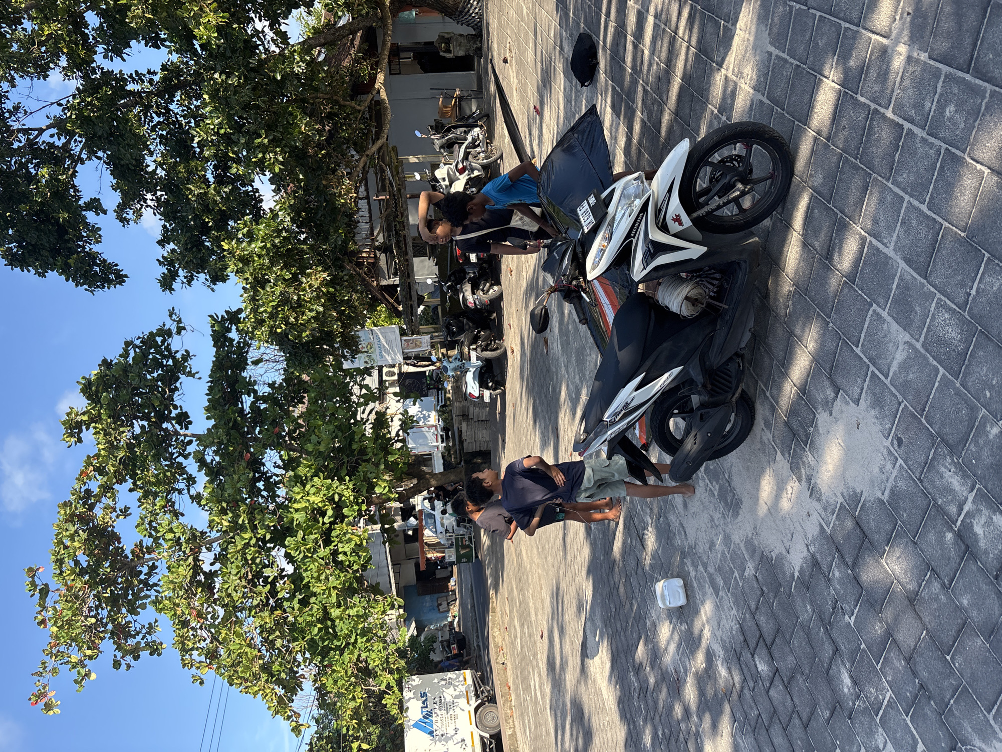 Two people are near a white and black Honda motorcycle. The photo appears to be taken in Kuta, Indonesia. The location shows paved brick parking with additional motorcycles parked in the background near shops. A mature tree casts shadows in the foreground and onto the pavement. Two people wearing casual clothes are standing on the brick pavers and looking at the motorcycle. There is a black motorcycle helmet resting on the motorcycle. A white plastic container sits on the ground near the feet of one person. The sky is visible through the tree foliage and is a light blue color.