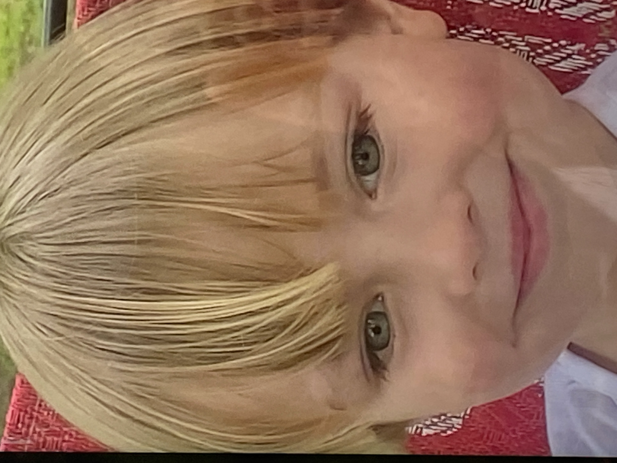 The image is a close-up portrait of a young child with blonde hair and blue eyes. The child is smiling slightly and looking directly at the camera. The background is blurred, but appears to be an outdoor setting with some greenery and a red textured surface. There is no specific information about the location in Pivdennoukrainsk, Ukraine, visible in the image.