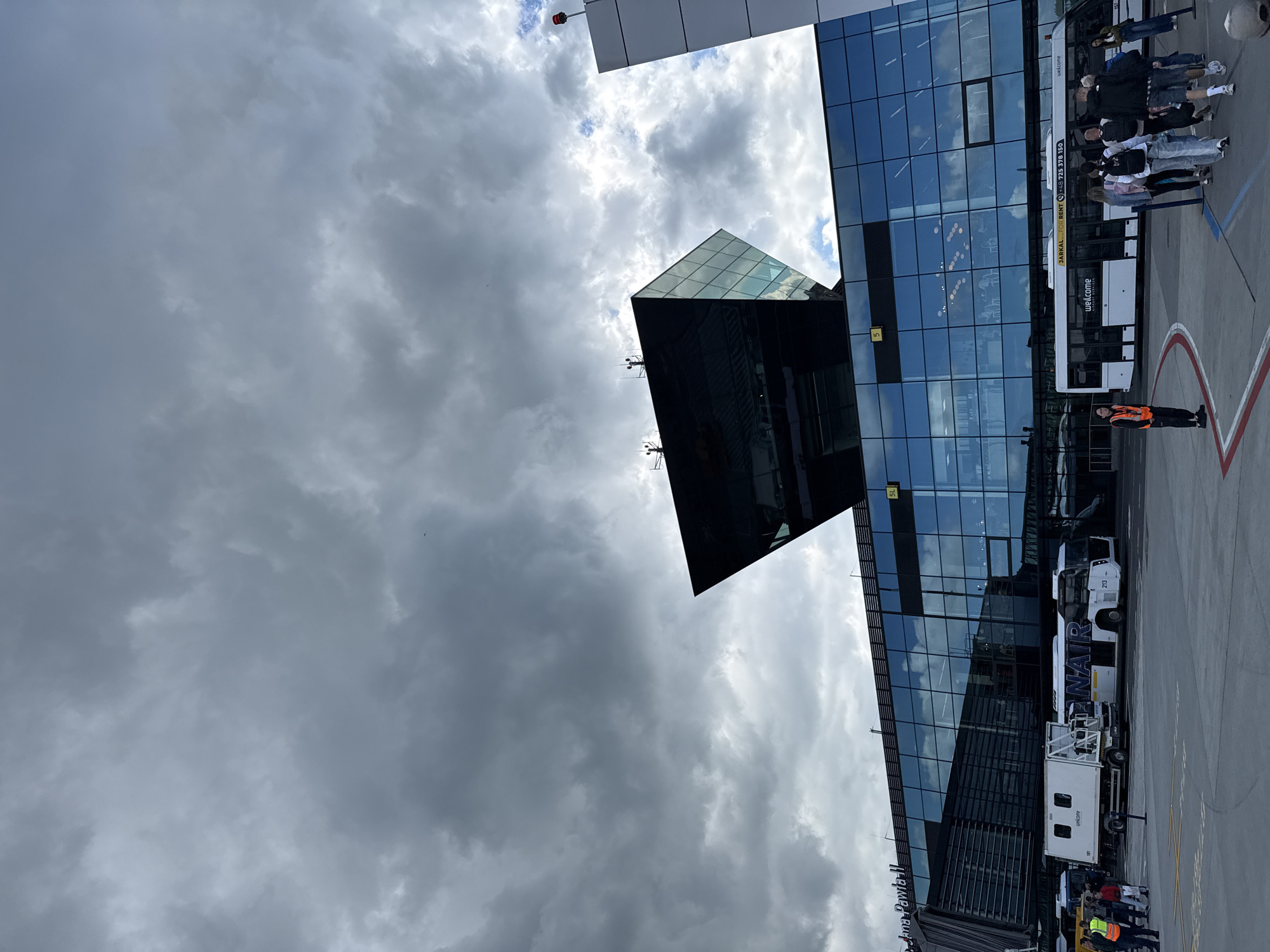 The image shows the exterior of John Paul II International Airport Kraków–Balice in Balice, Poland, under a heavily overcast sky. The modern airport terminal features a striking geometric glass and dark-colored facade. A distinctive, angular, dark glass structure sits atop the main building, resembling a control tower. Several buses are parked in front of the terminal, including one with "Ryanair" branding and another with "welcome" text. People are seen disembarking or waiting near the buses. A white service vehicle is also parked nearby. The overall scene depicts an active airport environment with a sense of impending weather change due to the dark clouds.
