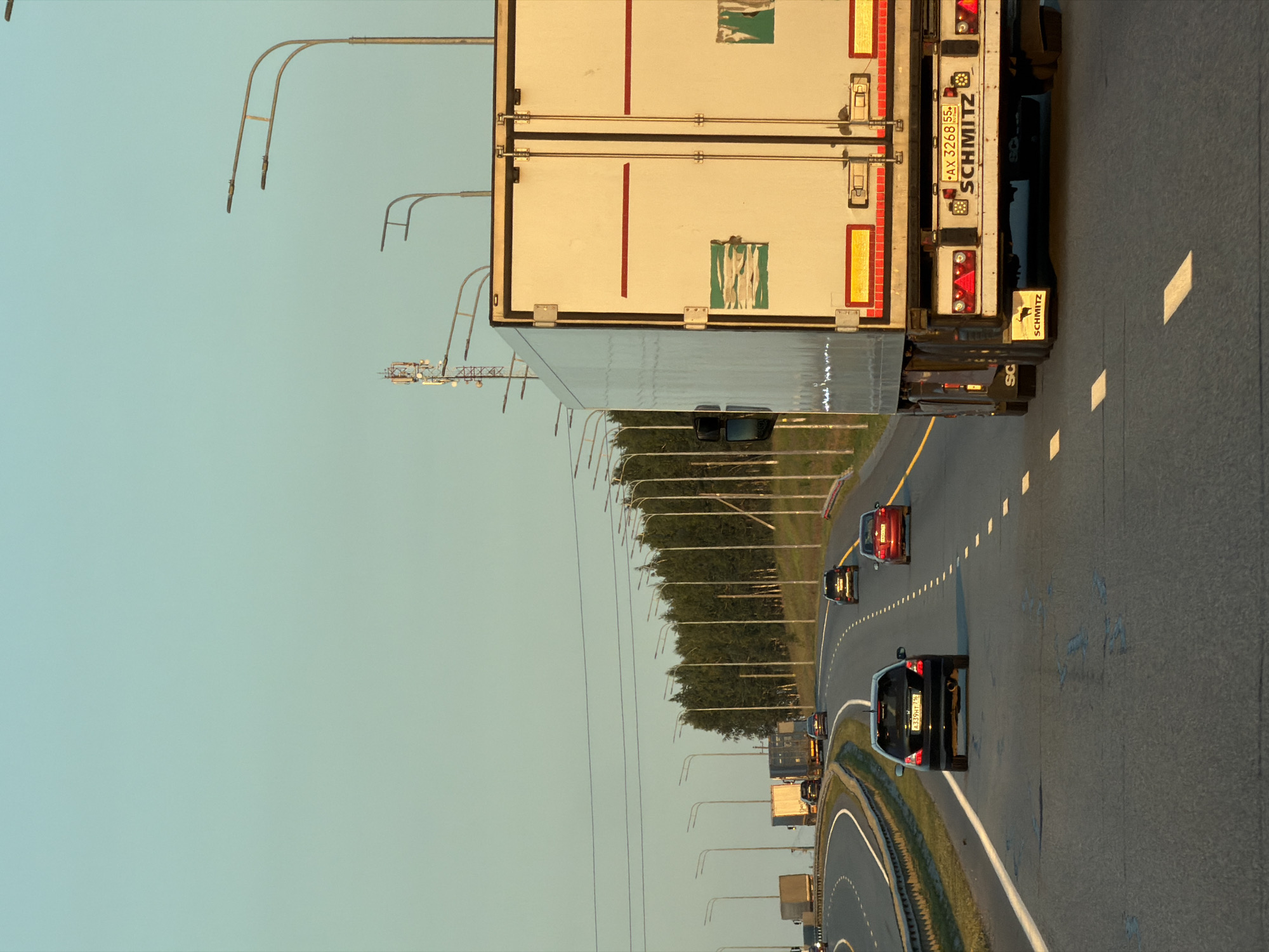 The image shows a highway with several vehicles, including trucks and cars. The perspective is from behind a large white truck in the foreground, which occupies a significant portion of the right side of the frame. This truck has a "SCHMITZ" logo on its rear. A black car is visible in the lane to the left of the foreground truck, with its license plate displaying "A339HT 77rus". Further down the highway, other vehicles, including a red car and a black SUV, are also traveling. The highway is flanked by a line of trees on the right and a series of streetlights on both sides. The sky is a clear, pale blue, suggesting it is either early morning or late afternoon. The road surface is asphalt with white lane markings. In the background, a telecommunications tower is visible.