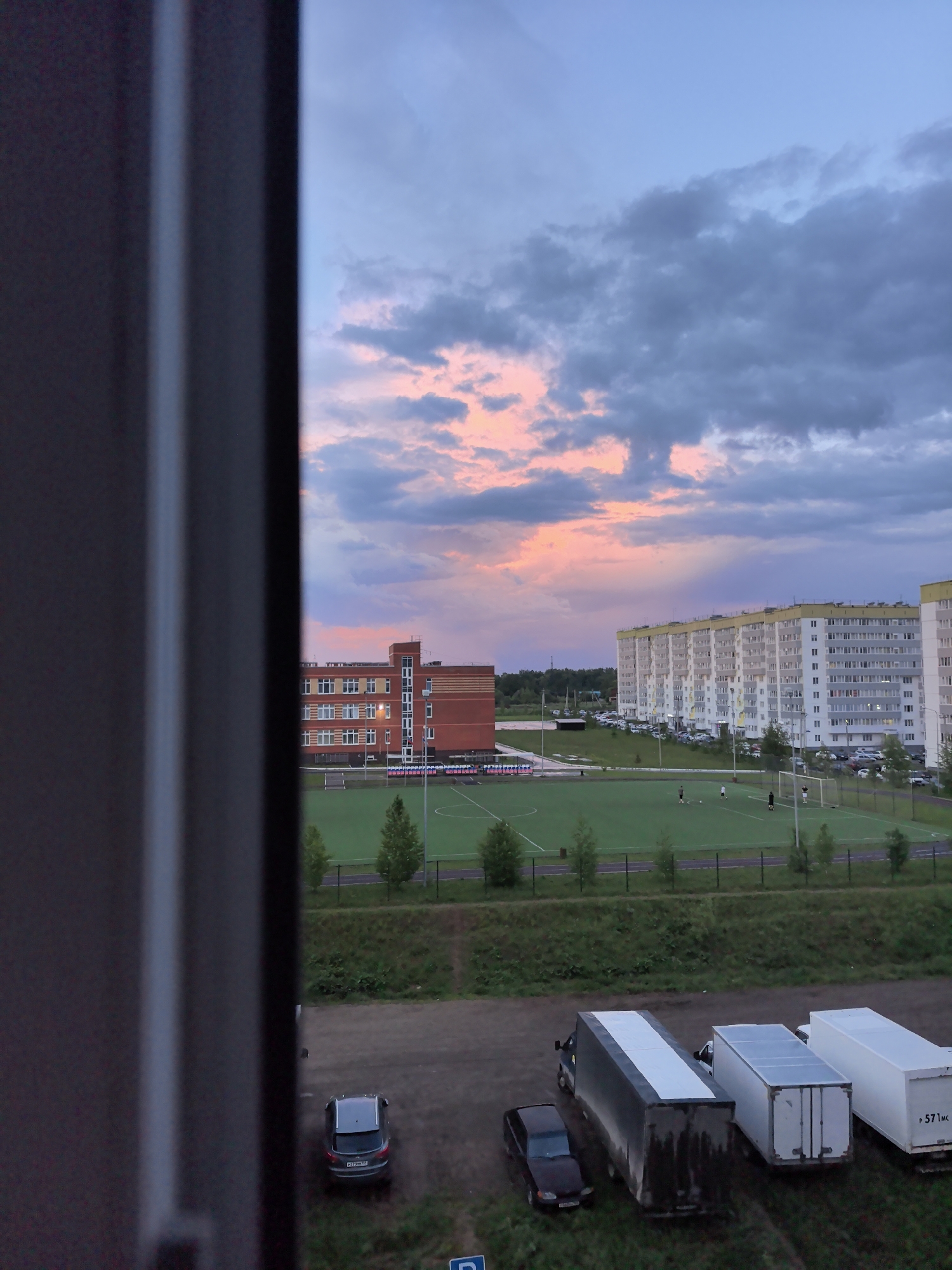 View from a window overlooking a soccer field, parking lot, and apartment buildings in Фролы, Russia. A school building is to the left of the field, and the buildings in the background are white with yellow trim. The sky is a mix of blue, grey, and pink clouds. There are several cars and trucks in the parking lot. People are playing soccer on the field.