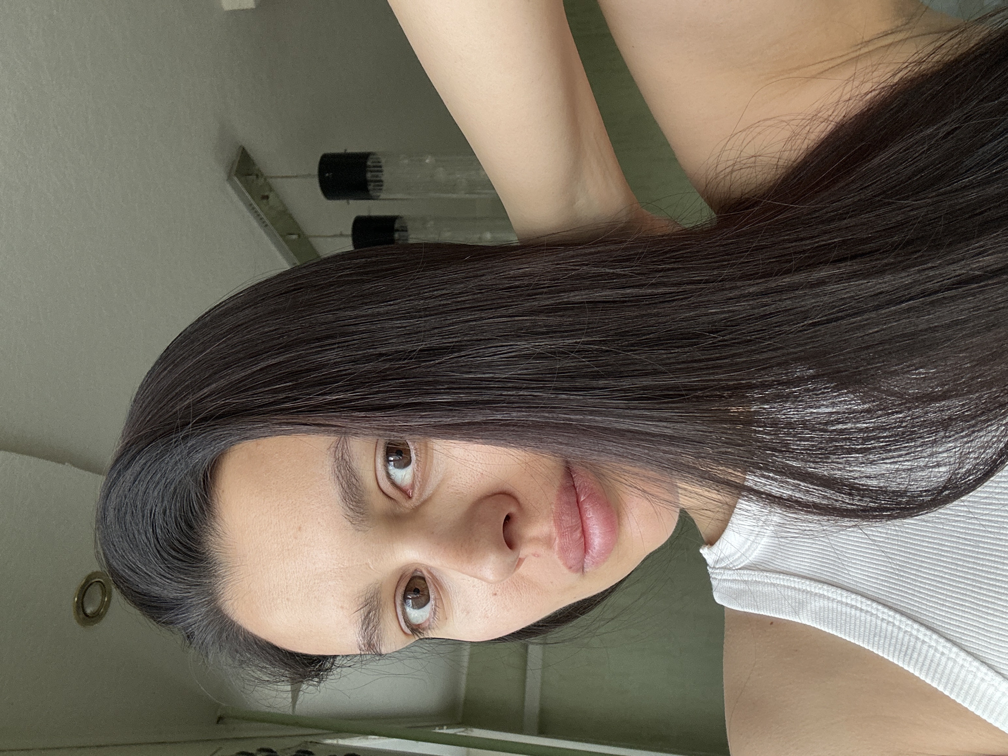 A close-up, angled selfie of a woman with long, dark brown hair. She has brown eyes, fair skin, and is wearing a white ribbed tank top. Her arm is raised, positioned behind her head. The background shows a ceiling with a light fixture and a hint of a wall. There is no way to place the image location in Chaikovsky, Russia from these details.