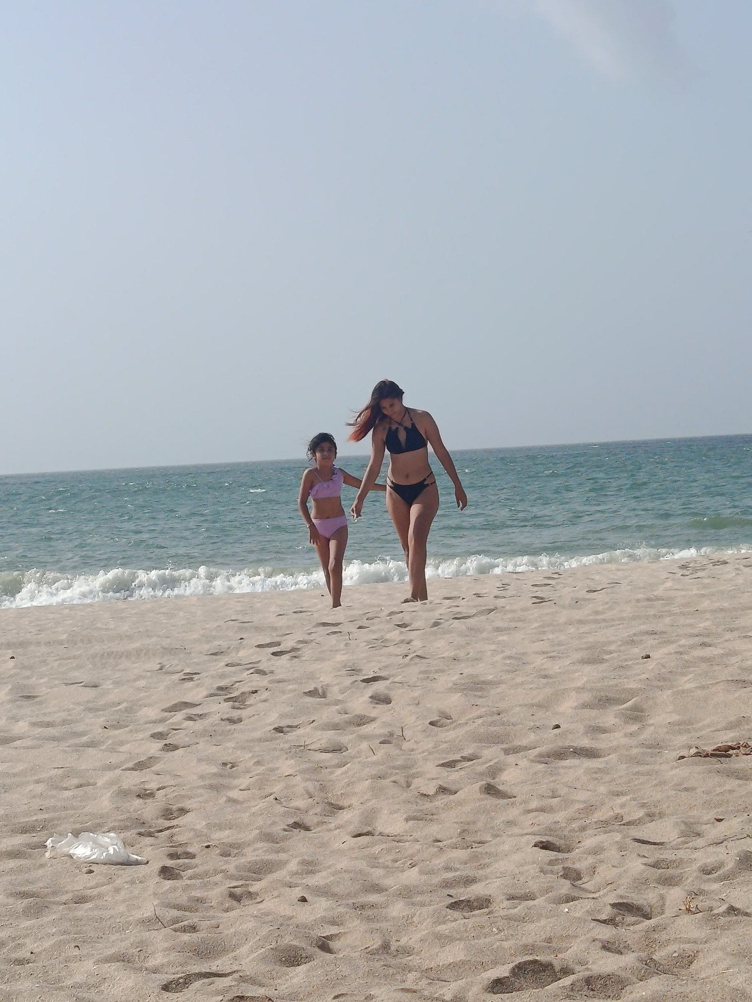 On a sandy beach in Pedro González, Venezuela, a woman and a child walk hand-in-hand. The woman is wearing a black bikini and the child is wearing a light purple bikini. The sea is in the background with gentle waves. There is some litter on the beach. The sky is light blue.