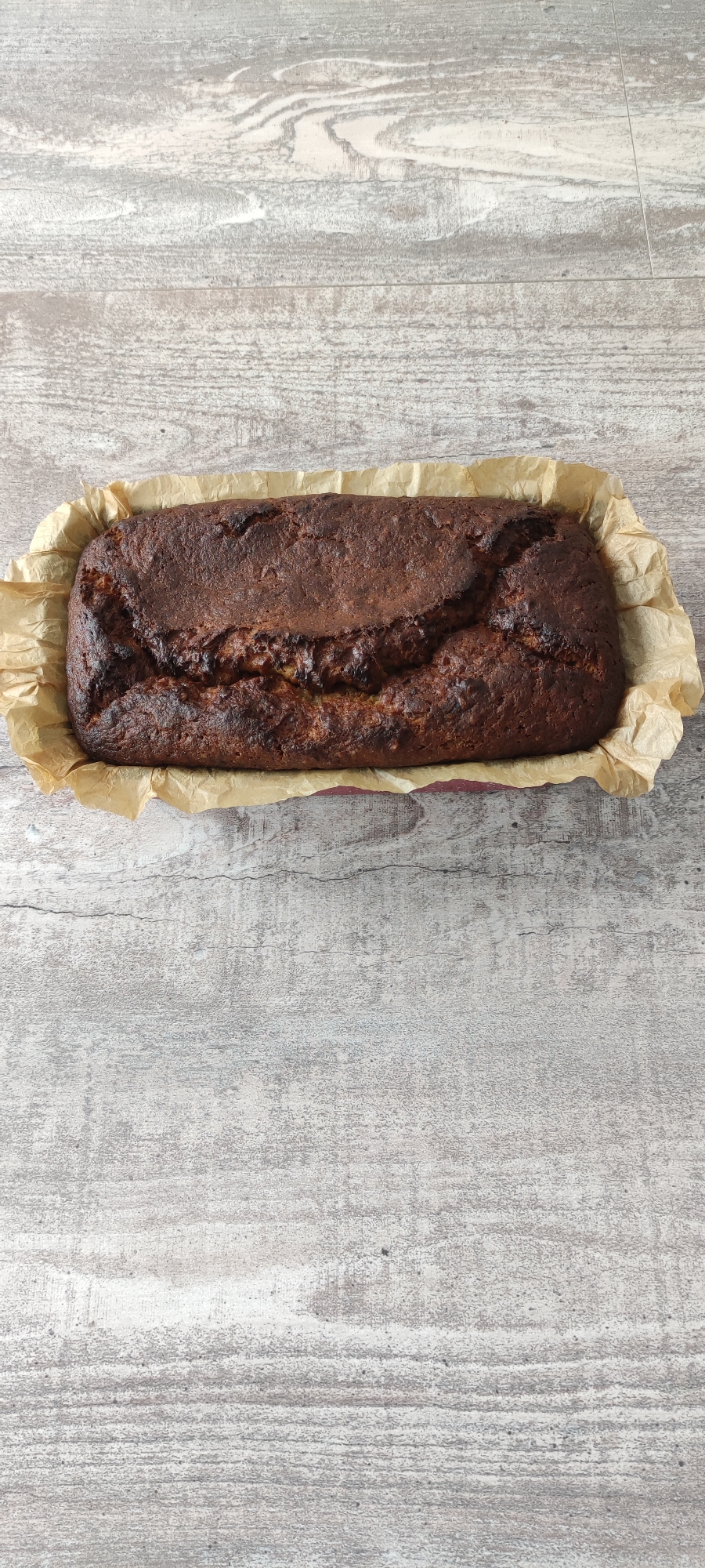 A close-up, overhead shot showcases a freshly baked loaf of bread, nestled within a rectangular red baking pan lined with crumpled parchment paper. The bread's surface is a deep, rich brown, with a pronounced crack running lengthwise down the center, revealing slightly lighter tones beneath. The loaf is positioned in the upper portion of the frame, against a backdrop of a light-colored, distressed wooden surface, exhibiting a weathered, planked appearance. The lighting is even, casting no distinct shadows, and the image is sharp, focusing solely on the baked good and its immediate surroundings. No people, actions, or interactions are depicted. The scene is devoid of any context that would place it in Rosinka, Russia, or any other specific location.