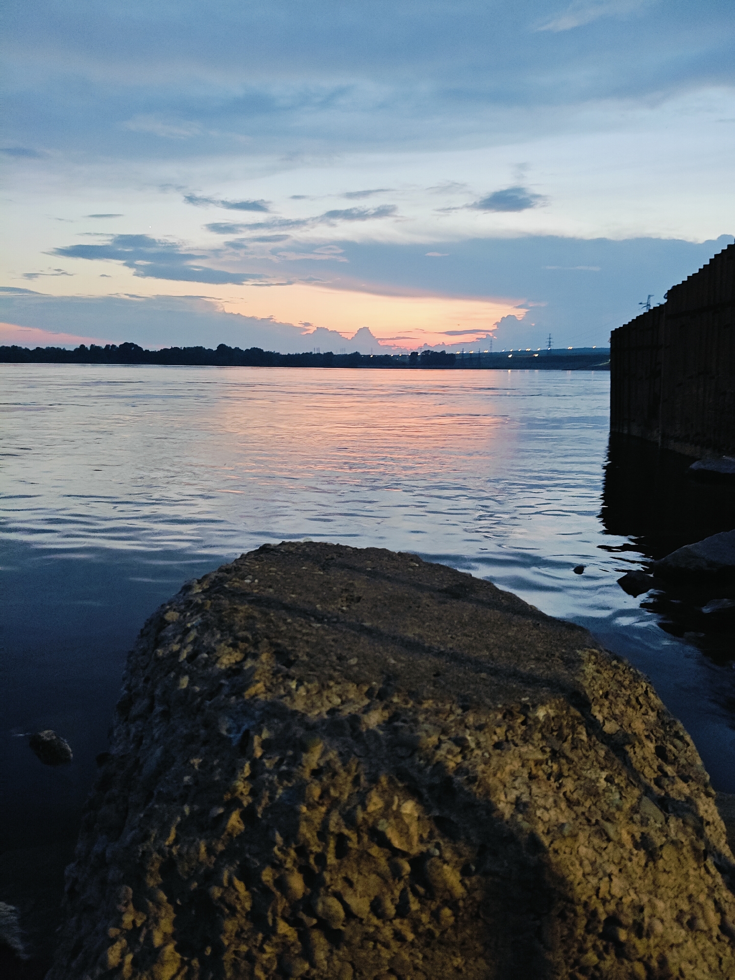 A wide shot captures a sunset over the Kama River near the city of Chaikovsky, Russia. The sky displays a gradient of blues and oranges, with scattered clouds reflecting the fading sunlight. A dark, rocky shoreline is in the foreground, with a large boulder taking up the lower right portion of the frame. The river's surface is calm, with gentle ripples reflecting the sky. In the distance, a silhouetted treeline borders the opposite bank, and industrial structures, possibly part of the Kama Hydroelectric Power Station, are visible on the right bank, marked by tall poles and lights. The scene is serene and evokes a sense of twilight.