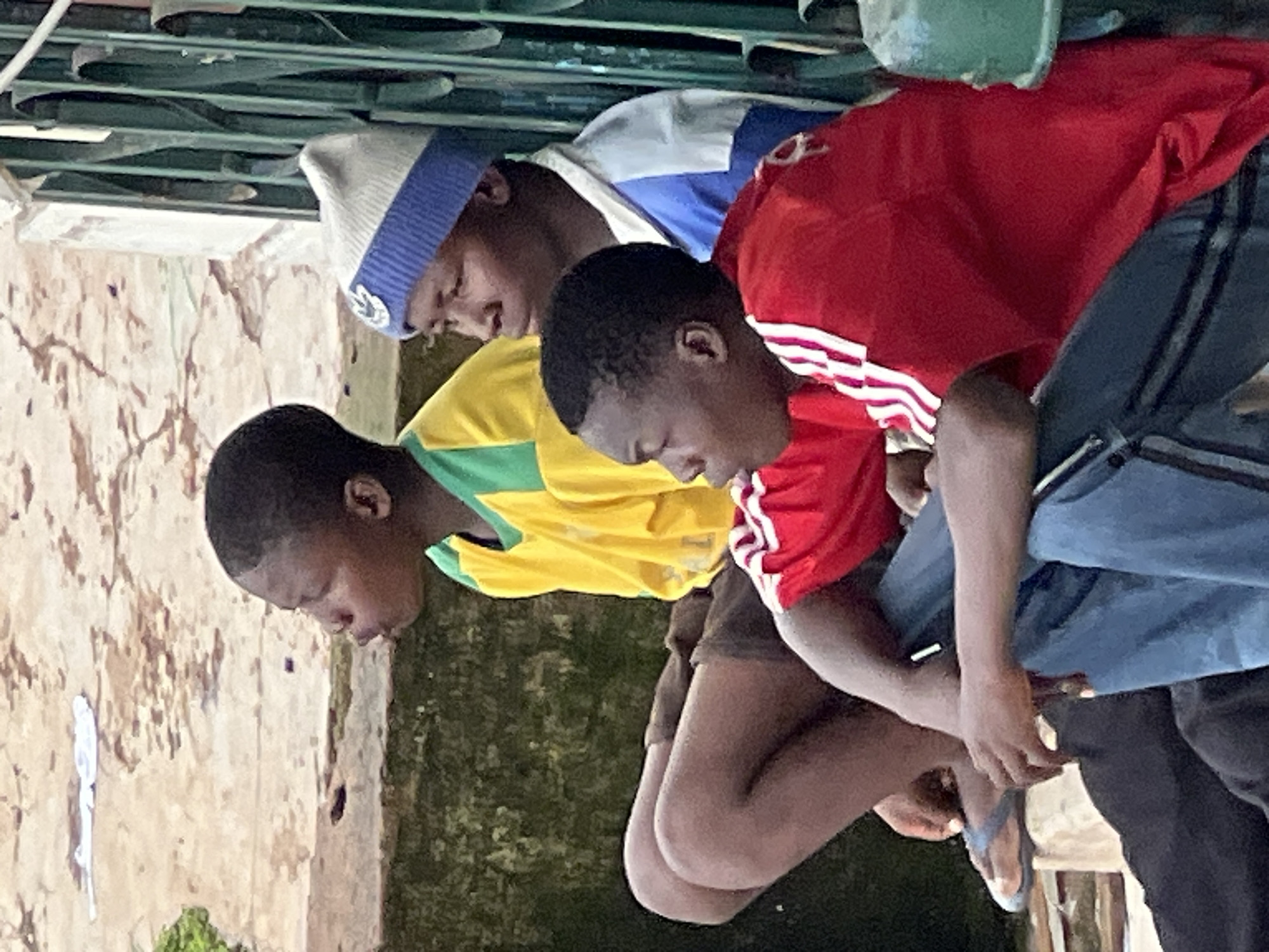 Three young Black males are seated outdoors, facing left. The foreground subject is wearing a red t-shirt with white stripes on the sleeves and blue pants. He is leaning forward, with his right arm resting on his right knee. The middle subject is wearing a yellow t-shirt with green trim around the collar. He is also leaning forward, with his knees bent. The background subject is wearing a blue and white t-shirt and a blue and white knit hat. He is looking to the left. They appear to be sitting on a concrete ledge, with a green metal fence behind them. The ground in front of them is covered in a mix of dirt and sand. The lighting suggests it is daytime. The scene is set in a general outdoor urban environment, possibly a residential area or public space, consistent with common settings in Jos, Nigeria.