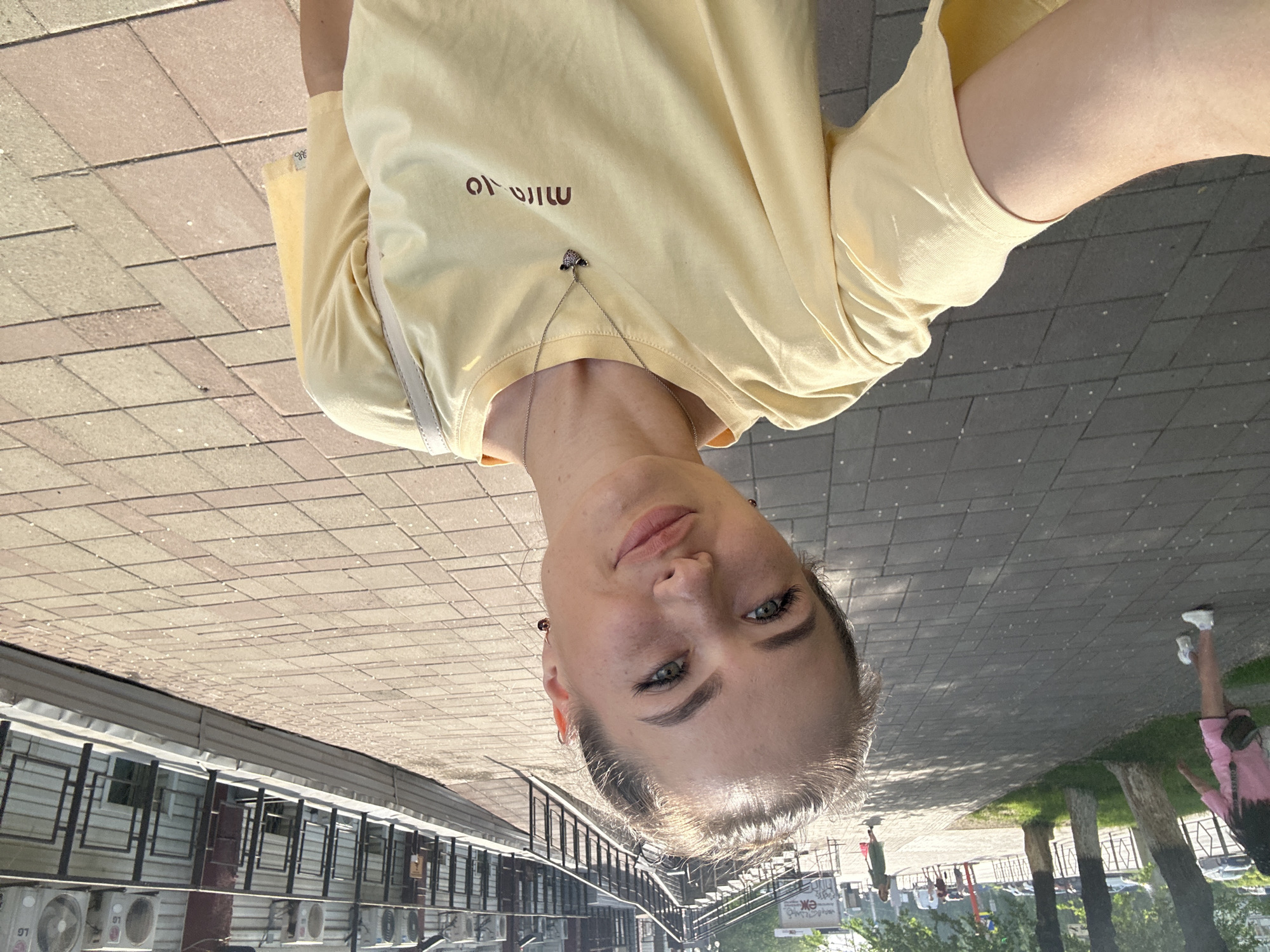 A young woman with blonde hair pulled back is taking a selfie outdoors on a sunny day. She is wearing a light yellow t-shirt and a necklace with a small charm. Her face is turned towards the camera with a slight smile. In the background, a paved walkway is visible, lined with trees on the left and a building with air conditioning units on the right. Other people and cars can be seen in the distance, suggesting an urban environment. The lighting suggests it is daytime, possibly late afternoon given the angle of the sun casting shadows.