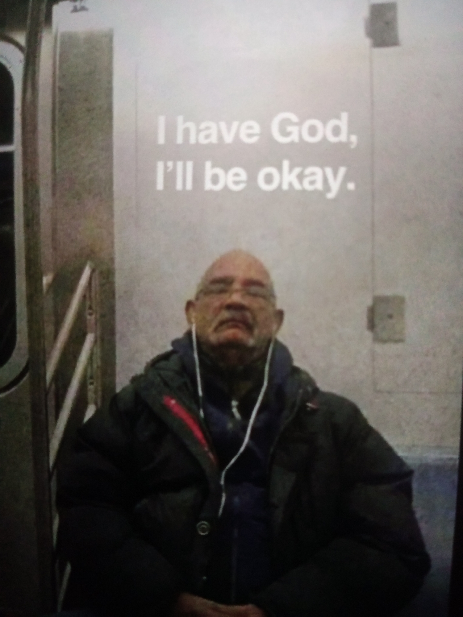 The image shows a man sitting on what appears to be a subway car. He has a bald head, is wearing glasses, and has earbuds in. He is wearing a dark jacket over a blue hooded sweatshirt. The background shows a wall with text that reads, "I have God, I'll be okay." The overall scene suggests a moment of quiet reflection or peace. There is no information in the image to suggest this location is in Jalingo, Nigeria; it appears to be a public transportation setting in a Western country.