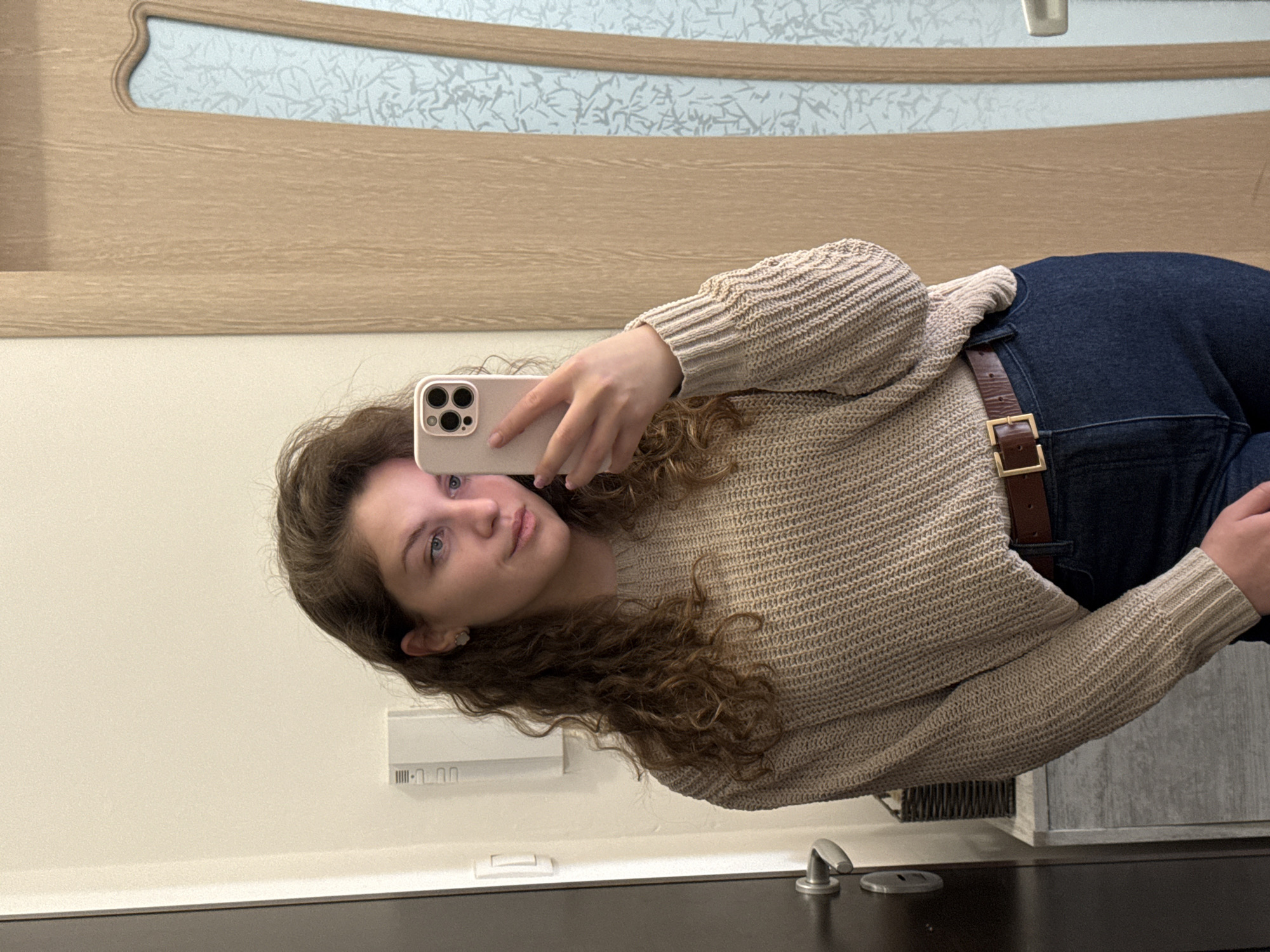 A woman is taking a selfie in a bathroom. She has curly brown hair and is wearing a tan sweater and blue jeans with a brown belt. The bathroom has a light-colored wall, a wooden door or paneling with a decorative insert, a dark countertop, and a modern faucet. The location is not identifiable as Sofia, Bulgaria based on the image alone.