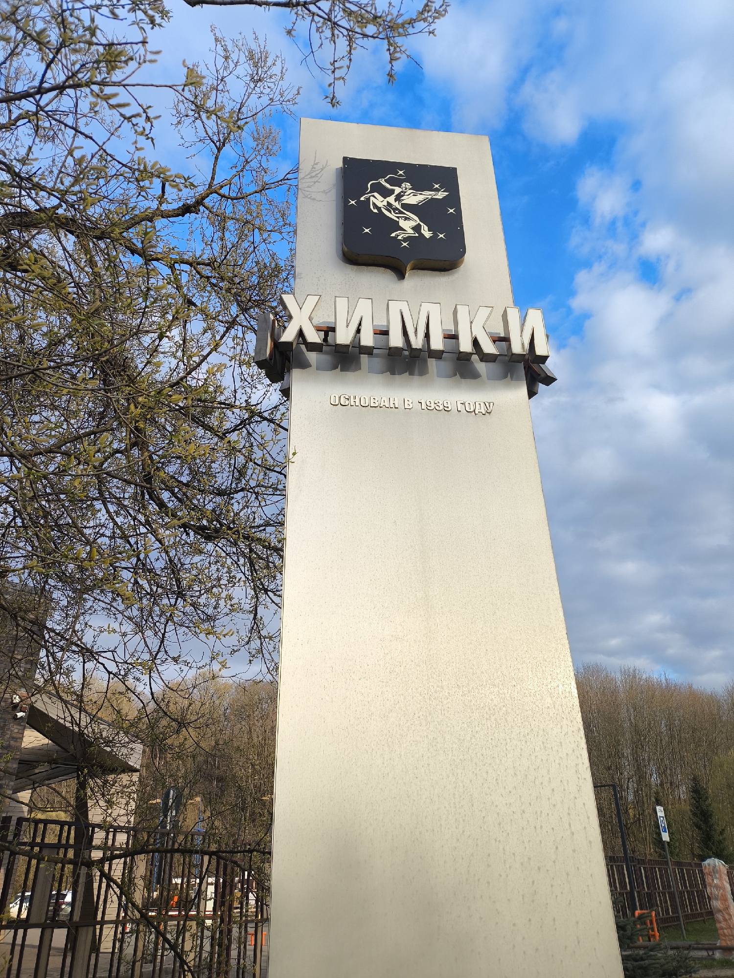 The image displays a tall, light-colored monument or sign, likely situated at an entrance or prominent location in a city. At the top of the monument is a dark coat of arms, featuring a stylized winged creature resembling a Pegasus, surrounded by stars. Below the emblem, large, three-dimensional metallic Cyrillic letters spell out "ХИМКИ" (Khimki). Further down, in smaller Cyrillic script, the text "ОСНОВАН В 1939 ГОДУ" (Founded in 1939) is visible.
The scene is set outdoors under a bright blue sky, partially covered by scattered white clouds, indicating a daytime shot with fair weather. On the left side of the frame, tree branches with light green budding leaves or catkins are prominent, suggesting springtime. These branches extend across the sky and partially obscure a distant building and a dark fence. On the right side of the monument, beyond a fence, more trees with emerging foliage are visible, along with a blue parking sign featuring a white "P" symbol. The monument itself appears to be made of a smooth, possibly metallic or concrete material.
There are no people or specific activities depicted in the image. It is a static shot primarily focusing on the informational sign. The location context indicates this is Khimki, Russia, which is directly supported by the visible text on the monument.