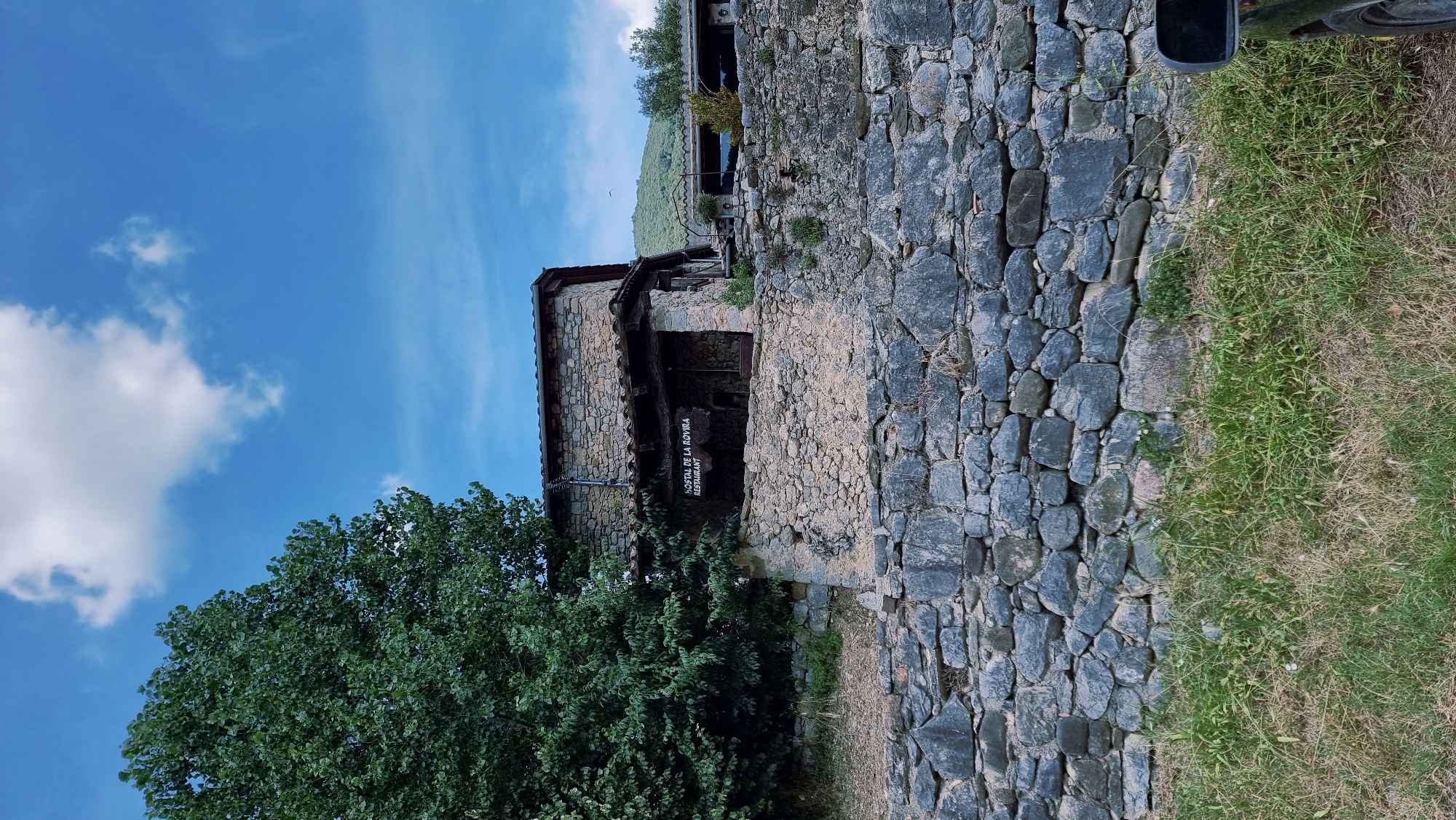 The image shows a rustic stone building with a sign that reads "HOSTAL DE LA ROVIRA RESTAURANT". The building appears to be a restaurant or an inn, likely located in a rural area given the surrounding stone walls, trees, and vegetation. A portion of a car mirror is visible in the lower right corner. The sky above is blue with scattered white clouds, and a green hillside is visible in the background. The overall scene suggests a peaceful and natural setting. The building is constructed from rough-hewn stones, with a tiled roof. A covered porch or veranda is visible in front of the main structure.