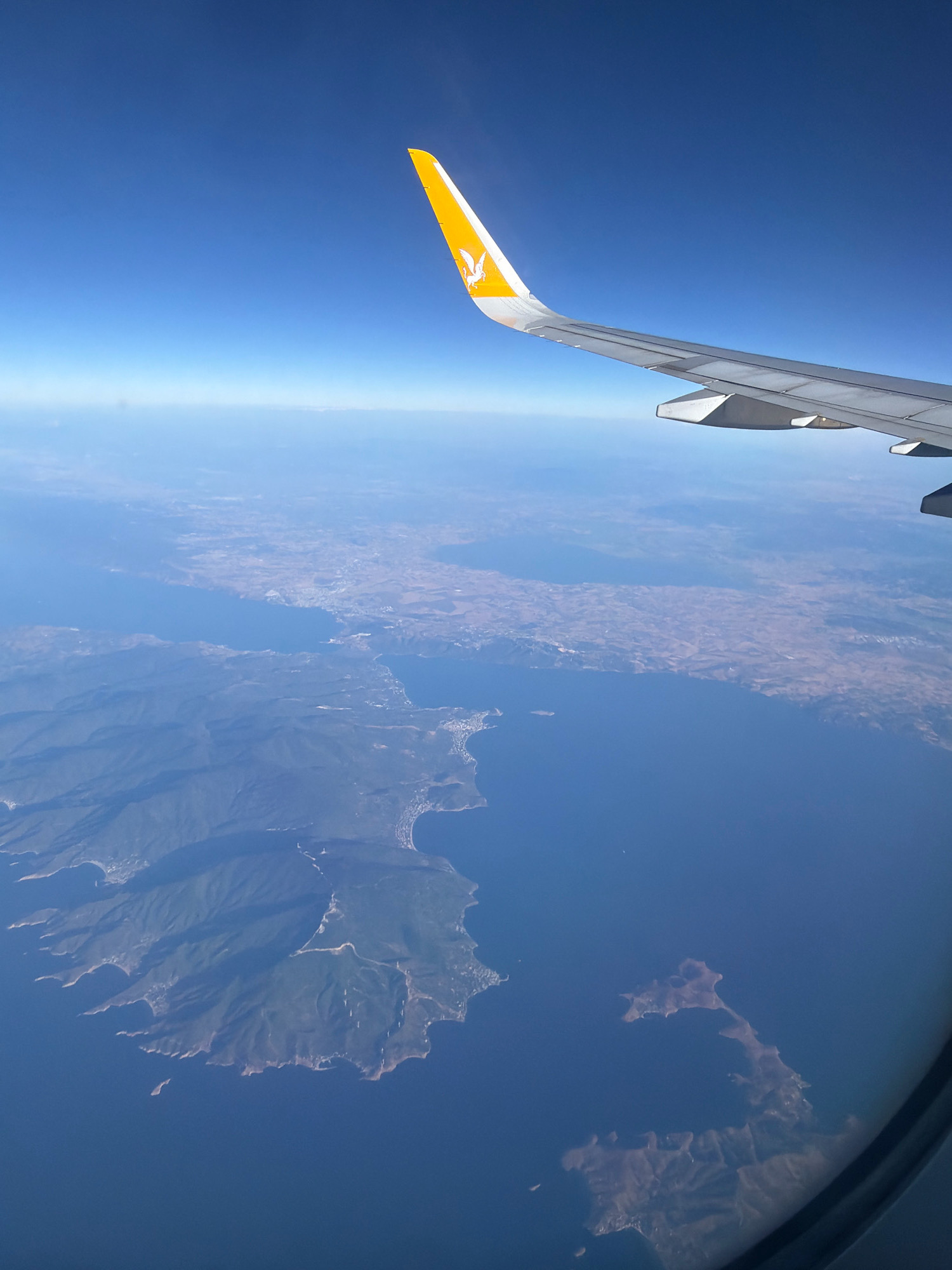 An aerial view from an airplane window showcases a vast expanse of deep blue sea meeting a rugged coastline. The wing of the airplane, with the Pegasus Airlines logo visible on its tip, extends into the upper right corner of the frame. The coastline is characterized by a mix of green, mountainous terrain and drier, flatter land, dotted with what appear to be settlements along the shore. Several peninsulas jut out into the sea, creating bays and inlets. In the distance, a hazy horizon separates the sea and land from the clear blue sky. One prominent island, visible in the lower middle of the frame, displays a winding coastline and some scattered structures. The overall impression is one of a scenic, coastal landscape viewed from a high altitude.