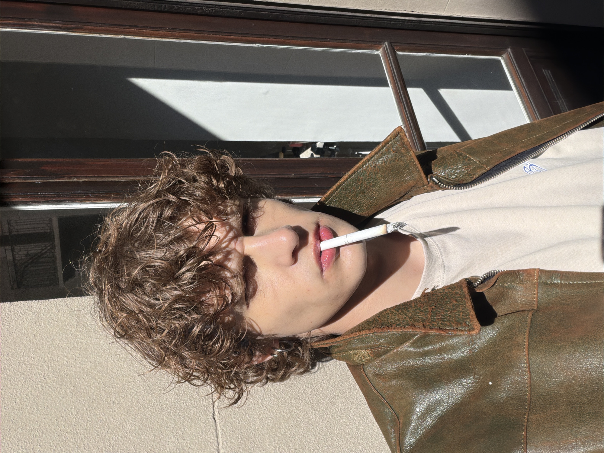 A young male individual is depicted in a close-up, outdoor setting in Milan, Italy. He possesses curly brown hair and light-toned skin, and a silver hoop earring is visible in his left ear. He wears a distressed olive-green leather jacket over a light cream t-shirt, which features a small blue embroidered logo on the left chest. A lit cigarette is positioned between his lips, with visible ash at its tip, indicating it has been partially consumed. His eyes appear partially shadowed, and his gaze is directed slightly downward. The background consists of a textured, light-colored building wall to the left of the subject. To his right, a dark brown wooden door or window frame with glass panes is visible. Strong sunlight illuminates the scene from the upper right, casting sharp shadows across the subject's face and creating a distinct diagonal shadow on the glass pane in the background.
