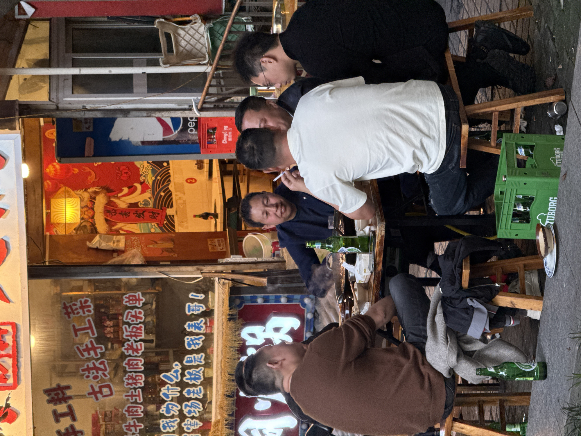 Here is a concise description of the image:
A group of men are sitting at a table outside an establishment, likely a restaurant, in the Yuzhong District, China. Some of them are drinking beer and smoking cigarettes. The restaurant has Chinese writing on the windows and signage, along with advertisements for Pepsi and Changying Beer. Empty bottles and a green crate of Tuborg beer are visible on the ground. The atmosphere is casual and relaxed.