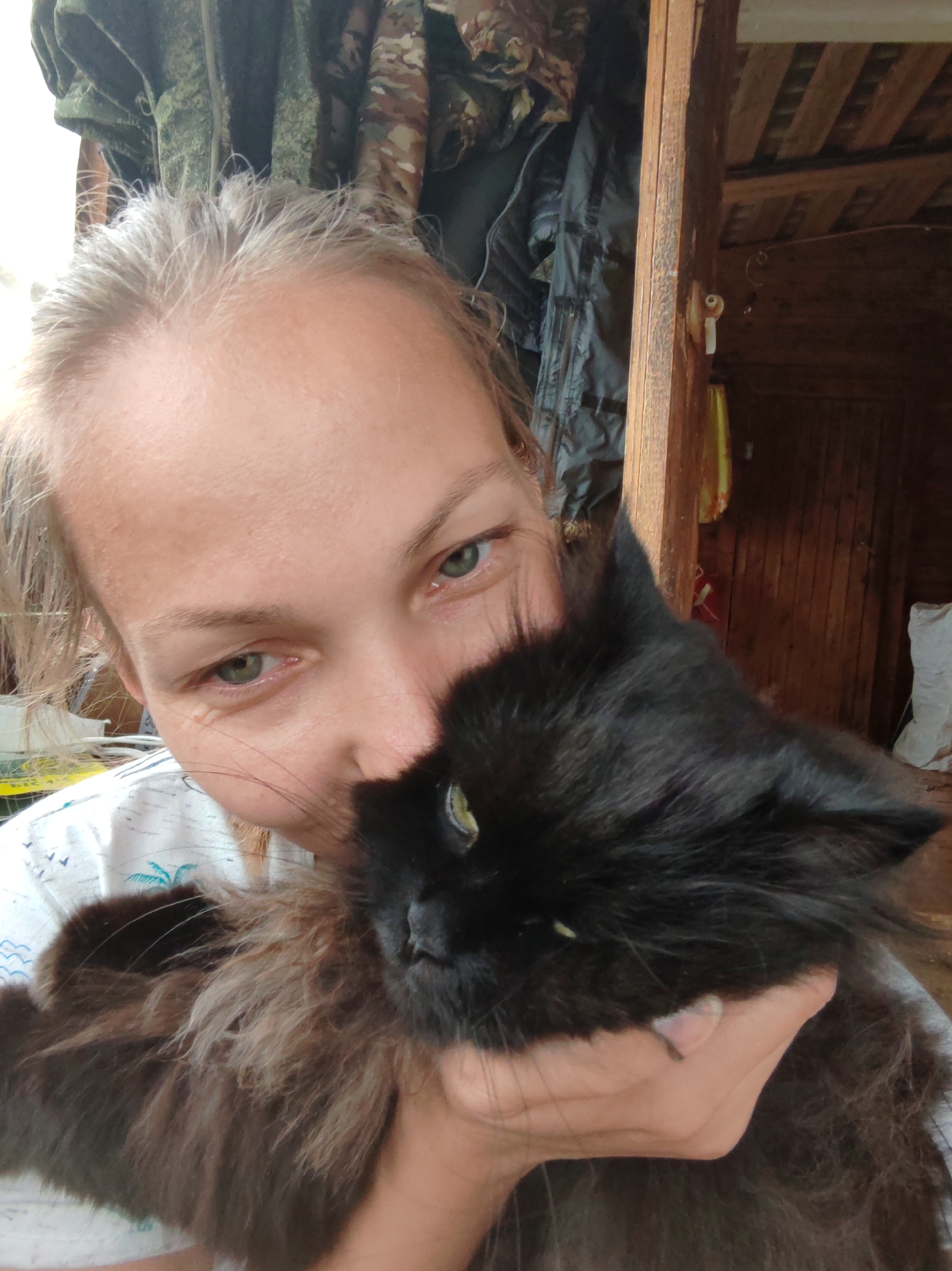A young woman with blonde hair is holding a black cat in her arms. She is looking at the camera with her eyes wide open. The cat is looking away from the camera. The background is blurred, but it appears to be a rustic interior, possibly a wooden house. There are some clothes hanging on the wall and a wooden beam. The image is taken from a close-up perspective, focusing on the woman and the cat. The overall impression is one of affection and warmth. The scene does not provide any specific details about the location in the city of Чухлэм, Russia.