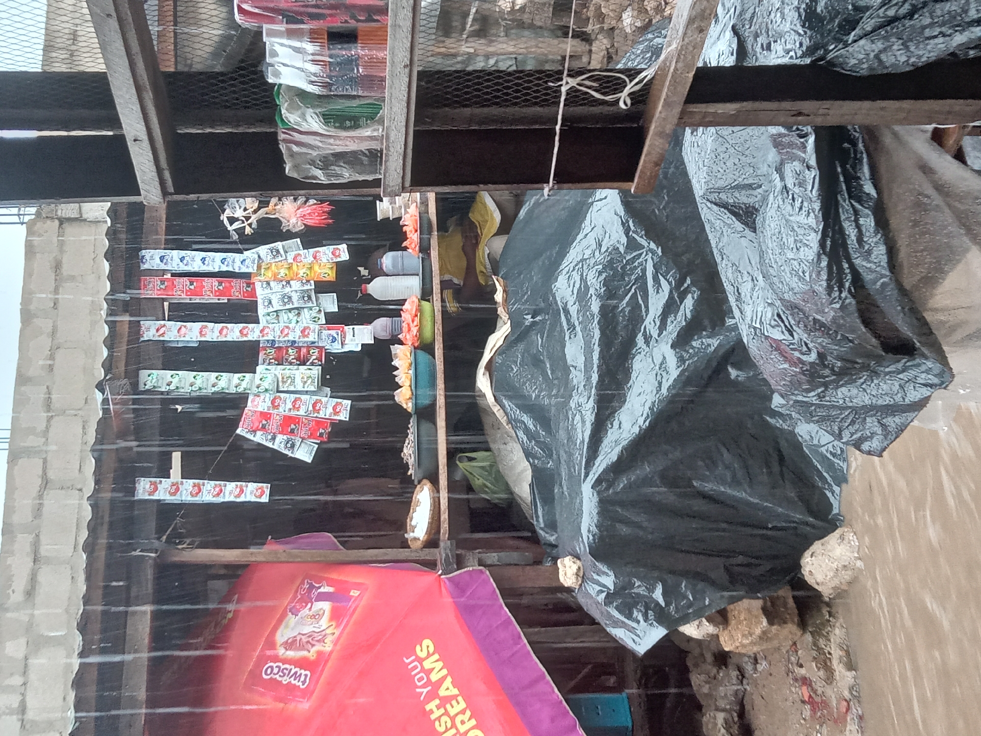 The image depicts a small shop in Sabon Tasha, Nigeria, possibly during rainfall. The shop is constructed of wood and corrugated metal, with a red awning bearing the "WISCO" logo and the phrase "WISH YOUR DREAMS". Merchandise, including small packets of goods and bottles, is displayed on shelves. A black plastic sheet covers part of the shop's structure. The surrounding area appears to be urban, with a cement block structure visible in the background. The ground is damp, suggesting rain. There are no people visible in the image.