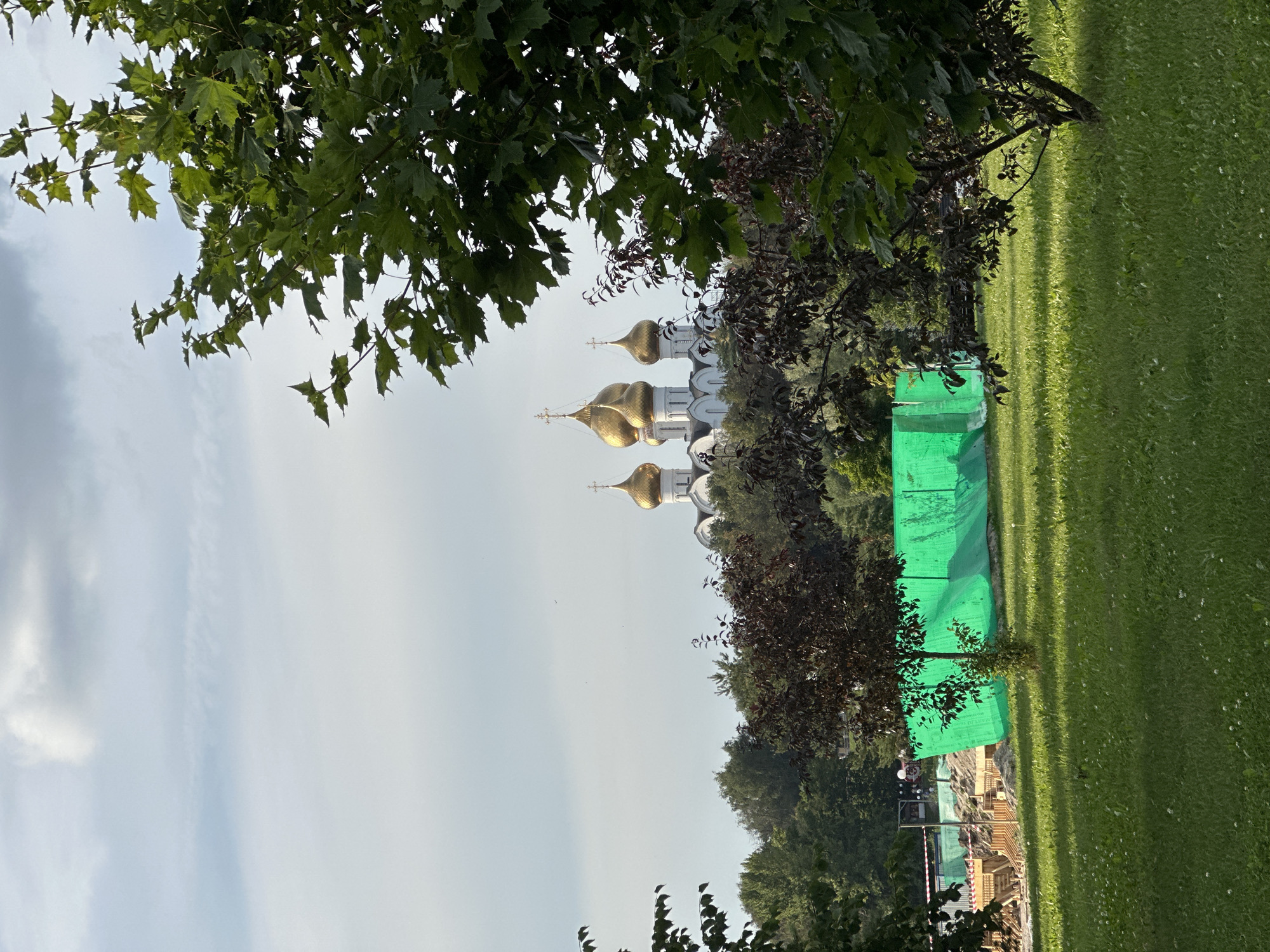 A low-angle, full outdoor shot captures a park area with a construction site in the mid-ground and a church with golden domes in the background. The foreground is dominated by a lush green lawn dotted with small white flowers. In the mid-ground, a construction site is partially obscured by a vibrant green mesh tarp. Wooden structures, possibly part of the construction, are visible behind the tarp. Several trees, one with dark foliage and another with lighter green leaves, stand in the mid-ground. In the background, atop a verdant hill, a white church with multiple golden domes and crosses is visible. The sky above is cloudy and light blue. The location is identified as Rusanovo, Russia.