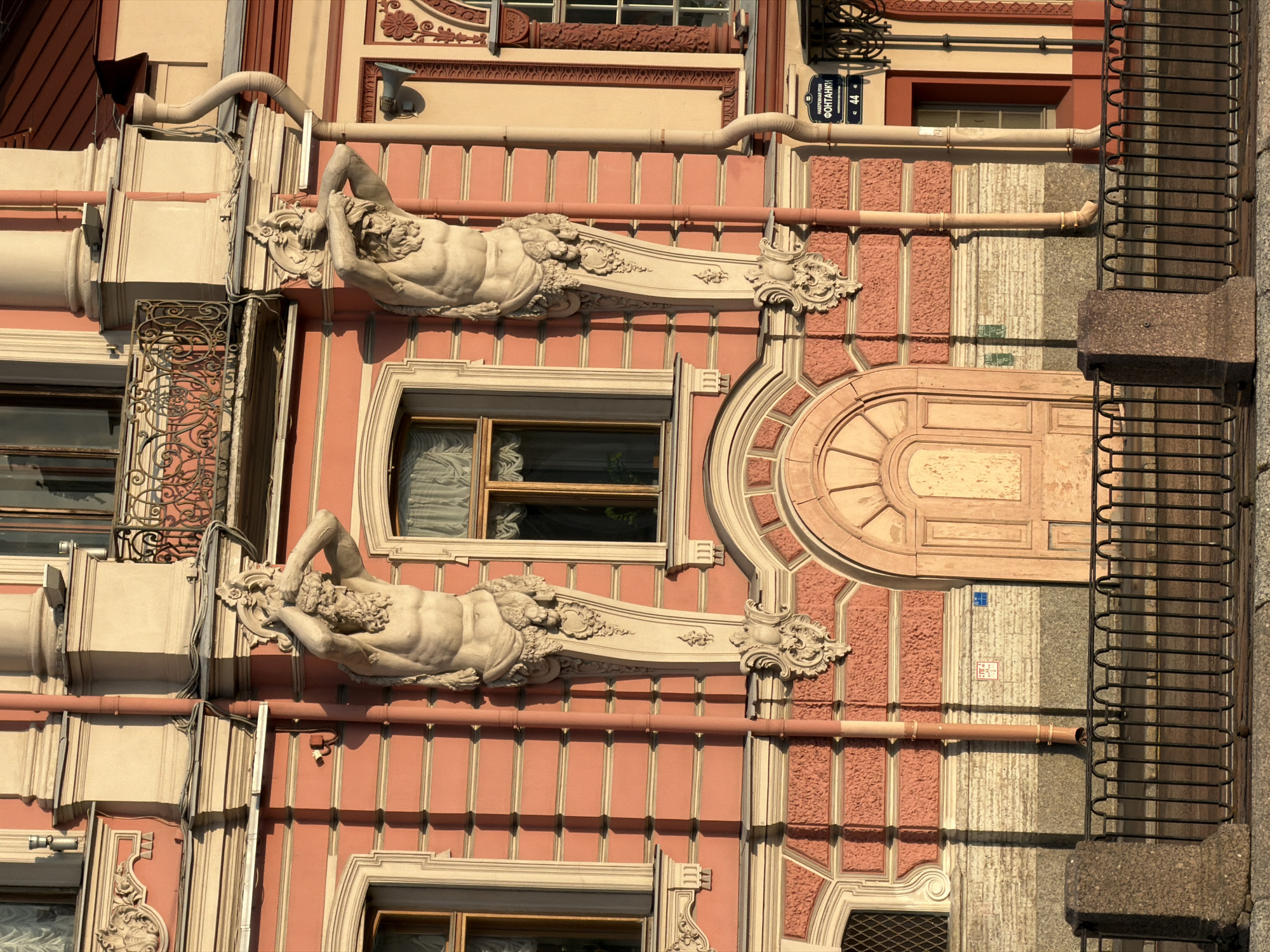 The image shows the facade of a building in Saint Petersburg, Russia, at the Fontanka River. The building is painted a salmon pink with beige decorative elements. Two large sculptures of muscular, bearded male figures, atlantes, are attached to the facade, supporting balconies or upper stories. These figures are carved in relief and appear to be made of stone. The building has several windows with wooden frames and curtains. A large arched wooden door is visible on the ground floor. A black metal fence runs along the bottom of the image, separating the building from what appears to be a sidewalk or embankment. The lighting suggests it is either early morning or late afternoon, casting long shadows. A street sign visible on the building reads "НАБЕРЕЖНАЯ РЕКИ ФОНТАНКИ" (Fontanka River Embankment).