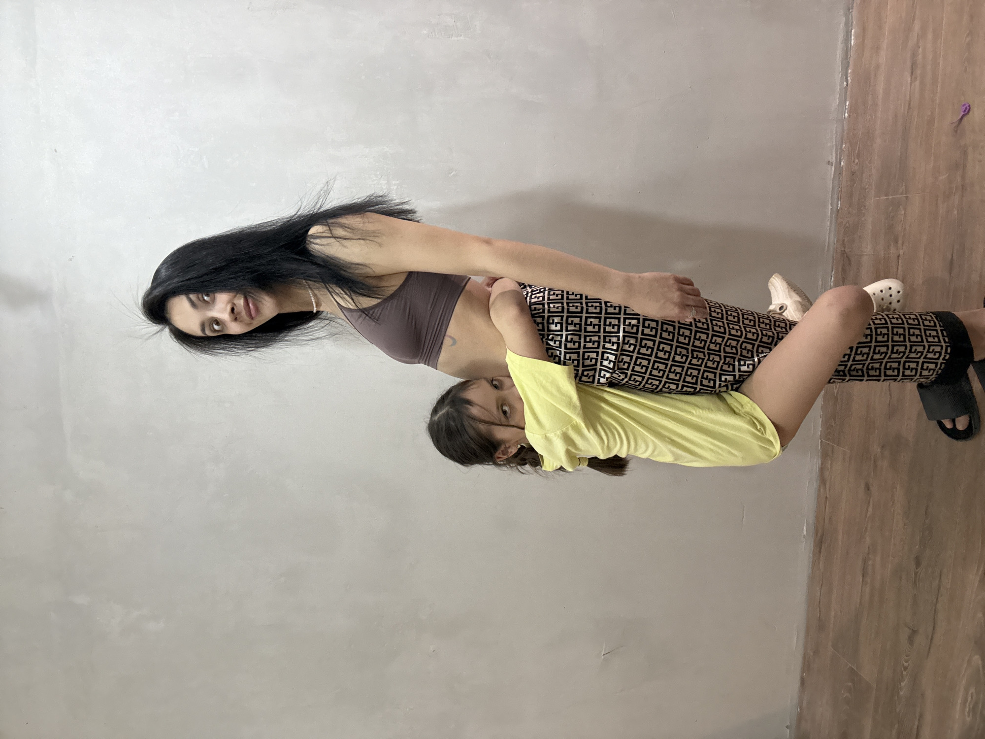 Two females are in a physical interaction. One female has long black hair and is wearing a brown sports bra, and patterned pants. The second female is wearing a yellow shirt. They are standing in a room with a gray wall and a wooden floor. The location is an indoor setting. Votkinsk, Russia cannot be inferred from the context of the image.