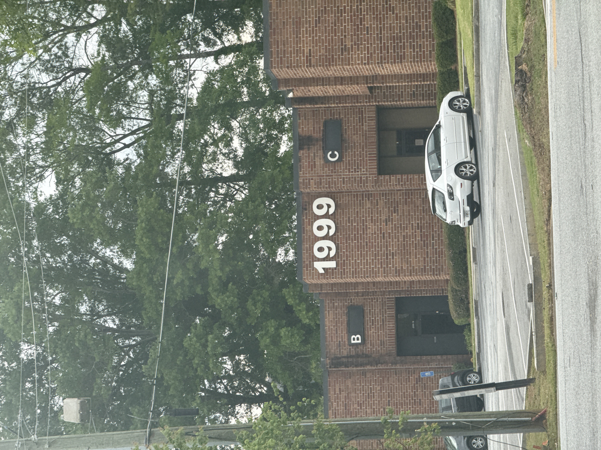 The image shows a brick building located at 1999, identified by large white numbers on the brick facade. The building has sections labeled "B" and "C". A white car is parked on the paved area adjacent to the building. The location is in Mountain Park, United States of America. A wooded area with tall trees is visible in the background. Overhead power lines are present. Another vehicle is partially visible at the bottom of the image. The scene appears to be on a cloudy day.