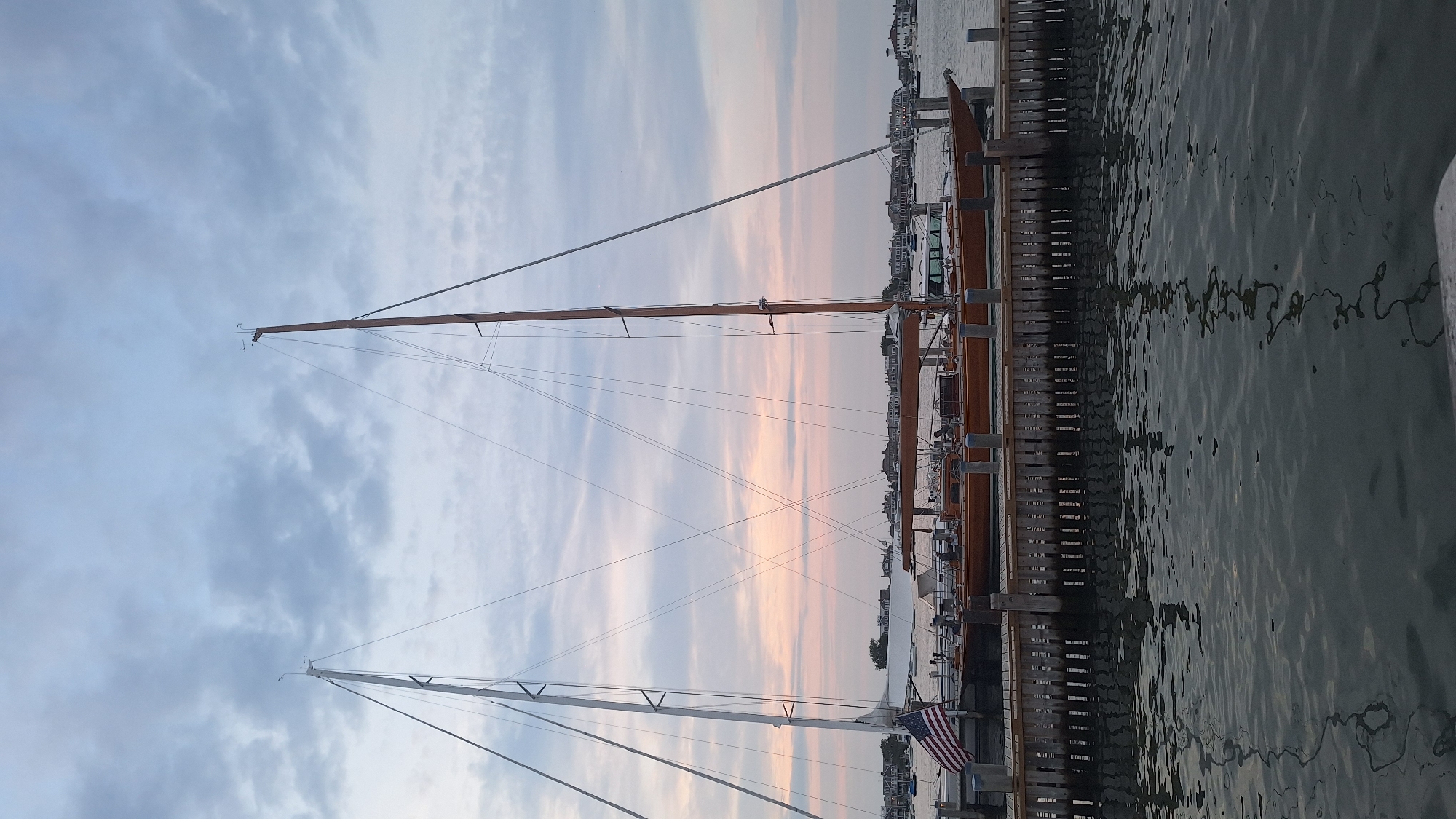 The image depicts a serene waterfront scene in Nantucket, United States of America, likely during a twilight hour given the soft pastel colors in the sky. Two prominent sailboats are docked side-by-side at a wooden pier. The boat on the left has a white sail partially unfurled and an American flag displayed. The boat on the right, with a wooden hull, has its mast clearly visible. The water in the foreground reflects the muted colors of the sky and the surrounding structures. In the background, a distant shoreline is dotted with houses, suggesting a residential or harbor area. The sky is filled with scattered clouds, adding texture and depth to the overall composition. The scene is peaceful and tranquil, capturing a quiet moment in a coastal town.