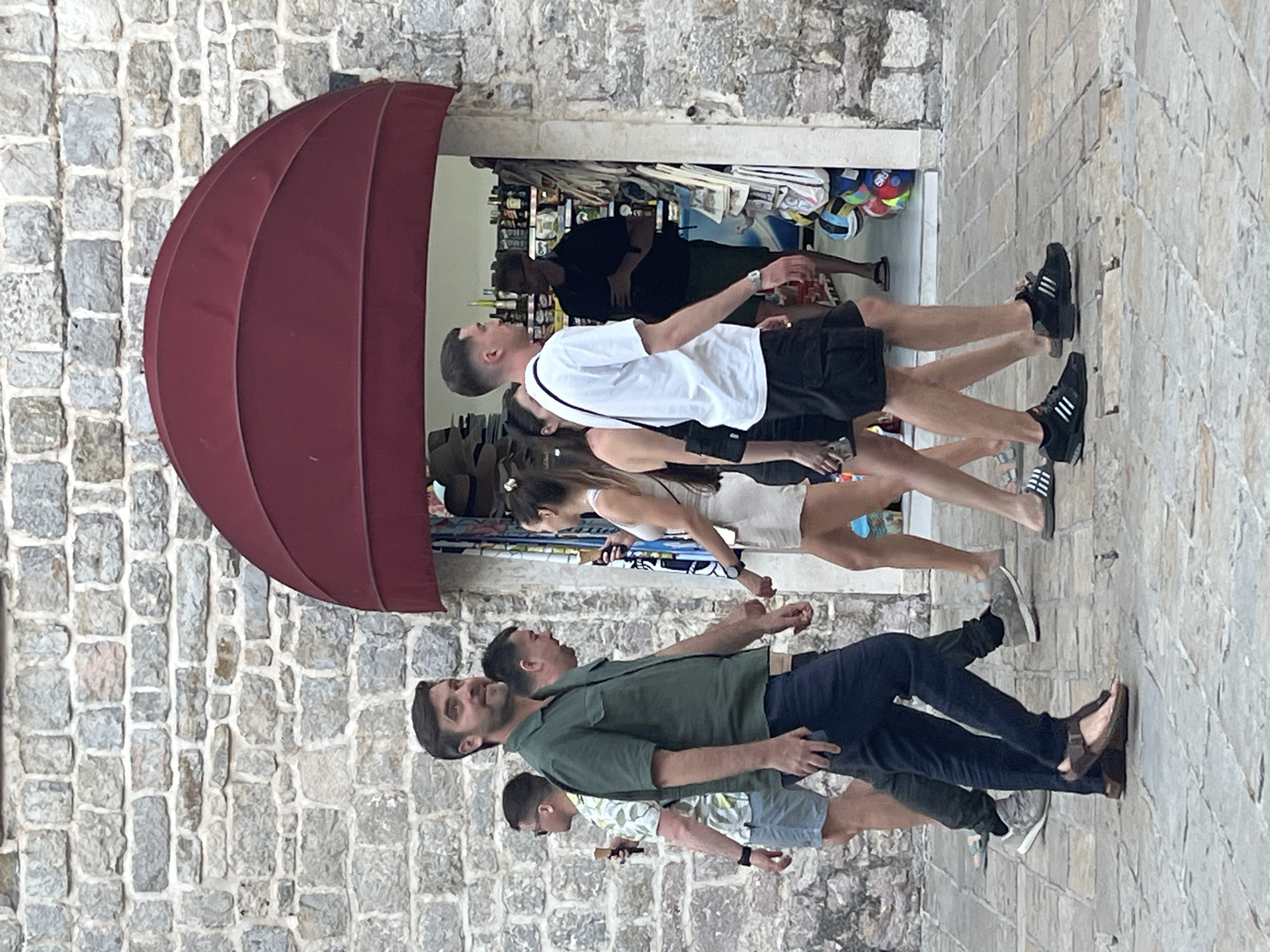 In Budva, Montenegro, a group of people walk along a stone-paved path in front of a building with a stone facade and a red awning over an open shop. The man in the foreground, wearing a green short-sleeved shirt and dark pants, looks to his left and smiles. To his left, a man in a patterned short-sleeved shirt and light shorts holds an ice cream cone and walks away from the camera. Behind the man in green, another man in a black shirt is partially visible. To the right of the man in green, a man in a white t-shirt and black shorts walks away from the camera. A woman in a light-colored dress and another woman in a dark top are walking ahead of him. Inside the shop, a man in a black t-shirt and green shorts stands behind a counter. The shop appears to be selling souvenirs or beach items. The overall scene depicts a casual, pedestrian street environment.