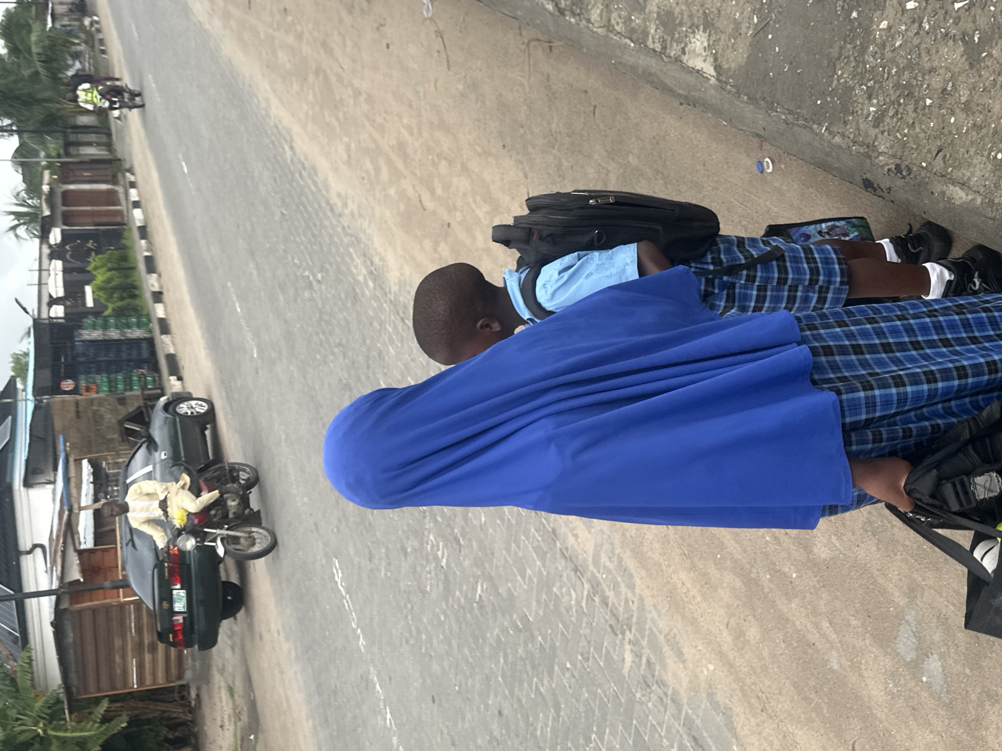 In Badagry, Nigeria, two children, a girl in a blue hijab and plaid skirt, and a boy in a blue shirt and plaid shorts, stand on a sidewalk beside a street. The girl holds a black bag. The boy carries a black backpack. In the background, a dark sedan is parked on the street, and a man in a white outfit rides a motorcycle. Further down the street, another person on a motorcycle is visible. Buildings and trees line the side of the road. The sidewalk is paved with light gray stones, and the street is asphalt.