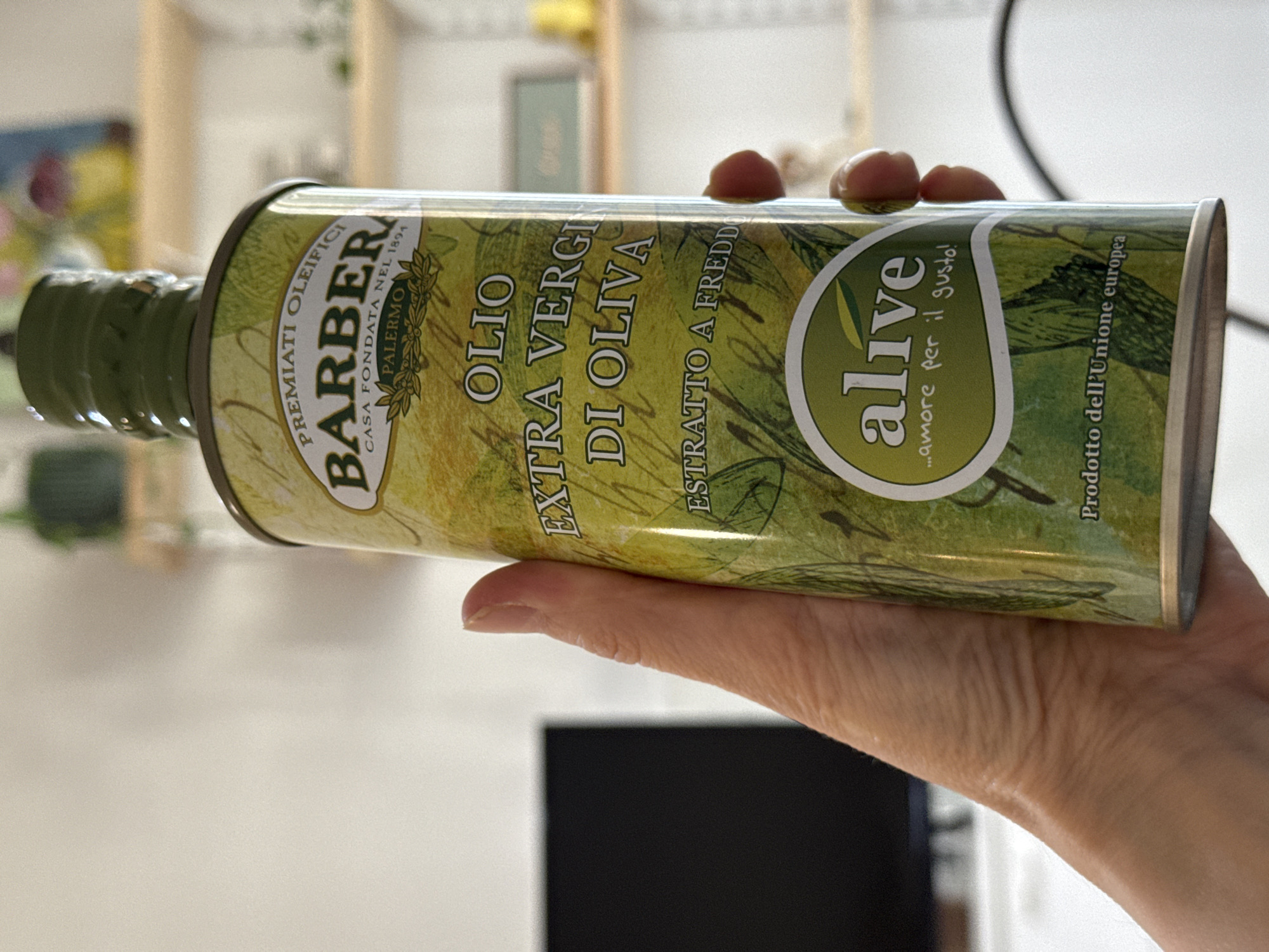 A hand is holding a horizontal cylindrical metal container of "BARBERA" brand olive oil, with a green plastic cap. The container's label indicates it contains "OLIO EXTRA VERGINE DI OLIVA," or extra virgin olive oil. The label also features the word "alive" in a stylized green circle, and includes the phrase "...amore per il gusto!" (Italian for "...love for taste!"). Text also identifies the product as "Prodotto dell'Unione europea" (Product of the European Union). The background is a blurred white wall with a light wood shelf unit with a few indistinct items.