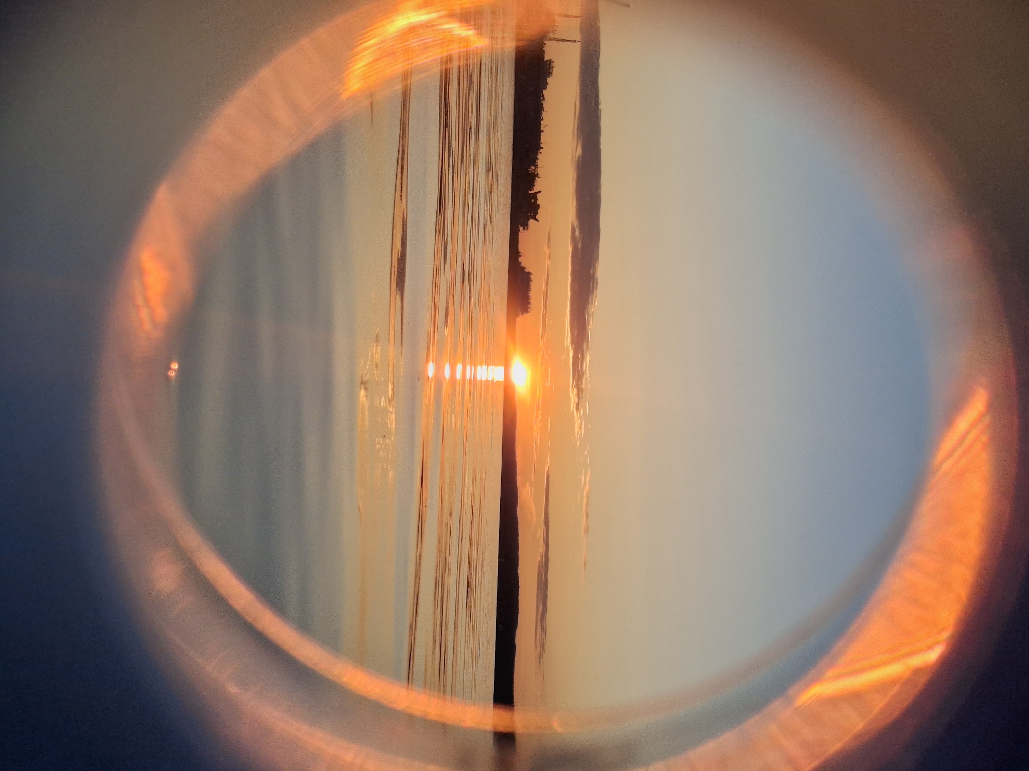 The image shows a sunset over a body of water, possibly the Kama River, as Chaikovsky is located on its banks. The sun's reflection creates a bright streak across the water's surface. Land with vegetation is visible along the shoreline. The scene is viewed through a lens or filter, creating a circular orange and yellow halo effect around the image. The foreground appears dark, suggesting the photograph was taken from within a shaded or darker area.