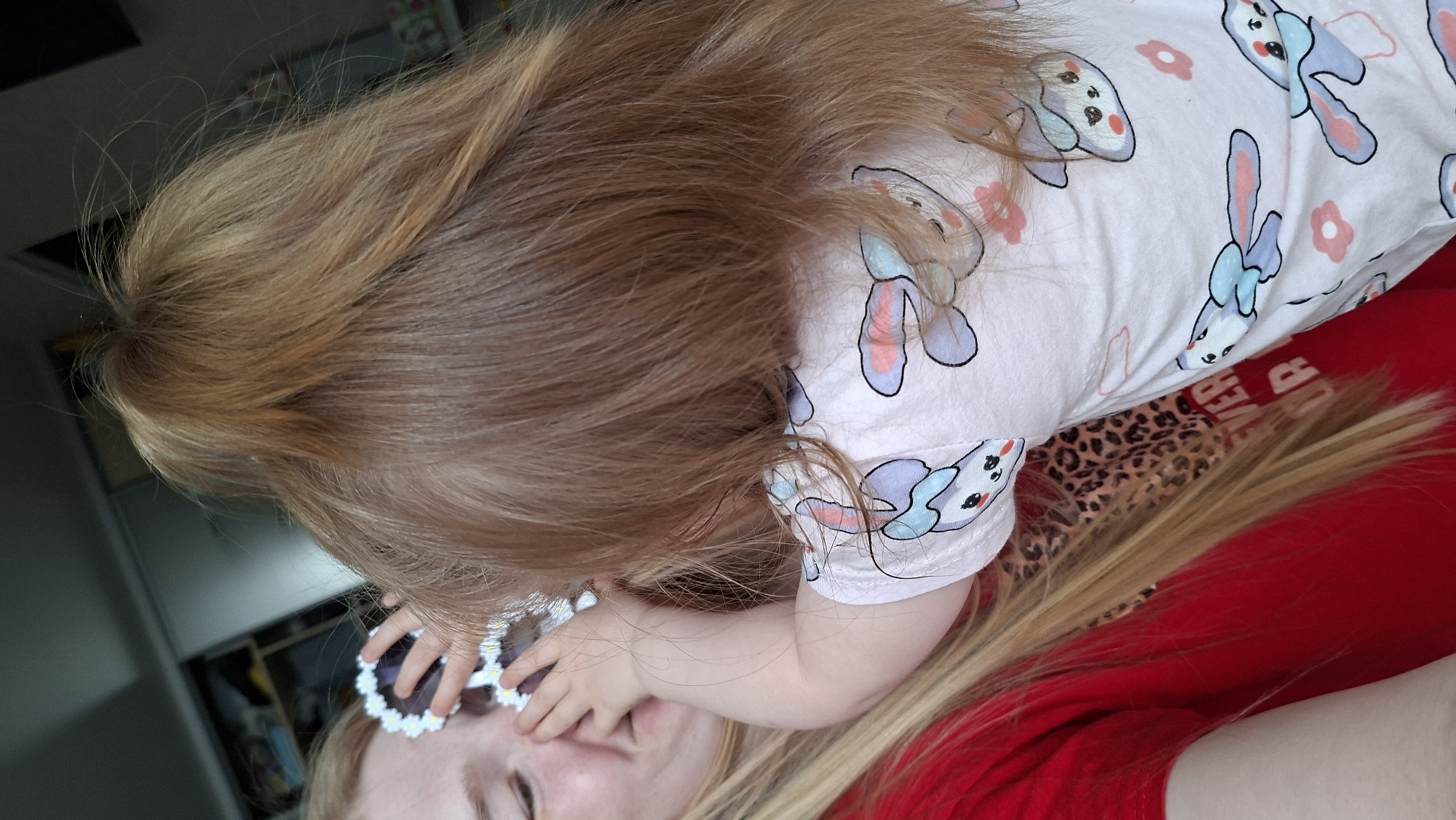 A young child is positioned above an adult, possibly their mother. The child has light brown hair and is wearing a light pink onesie with a bunny pattern. The child's hand is near the adult's face, and they are holding a pair of flower-shaped sunglasses. The adult is wearing a red shirt. The background appears to be indoors, with a glimpse of a cabinet or shelving unit. No information is available to determine if the location is in Chaikovsky, Russia.