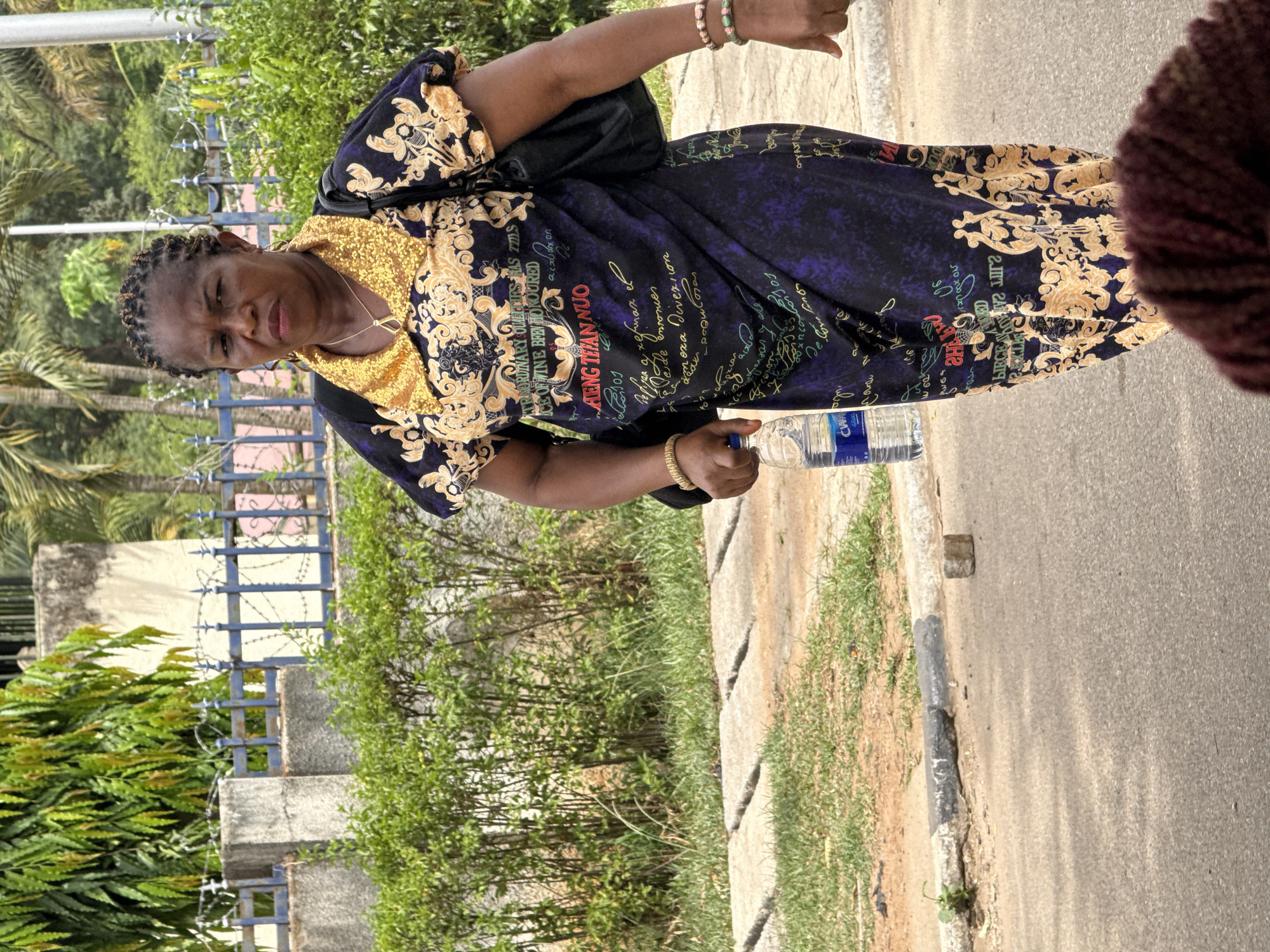 A woman in a blue and gold patterned dress stands on a sidewalk in Abuja, Nigeria, holding a bottle of water. A black bag is slung over her shoulder. The background includes green vegetation, a fence, and a paved road.
