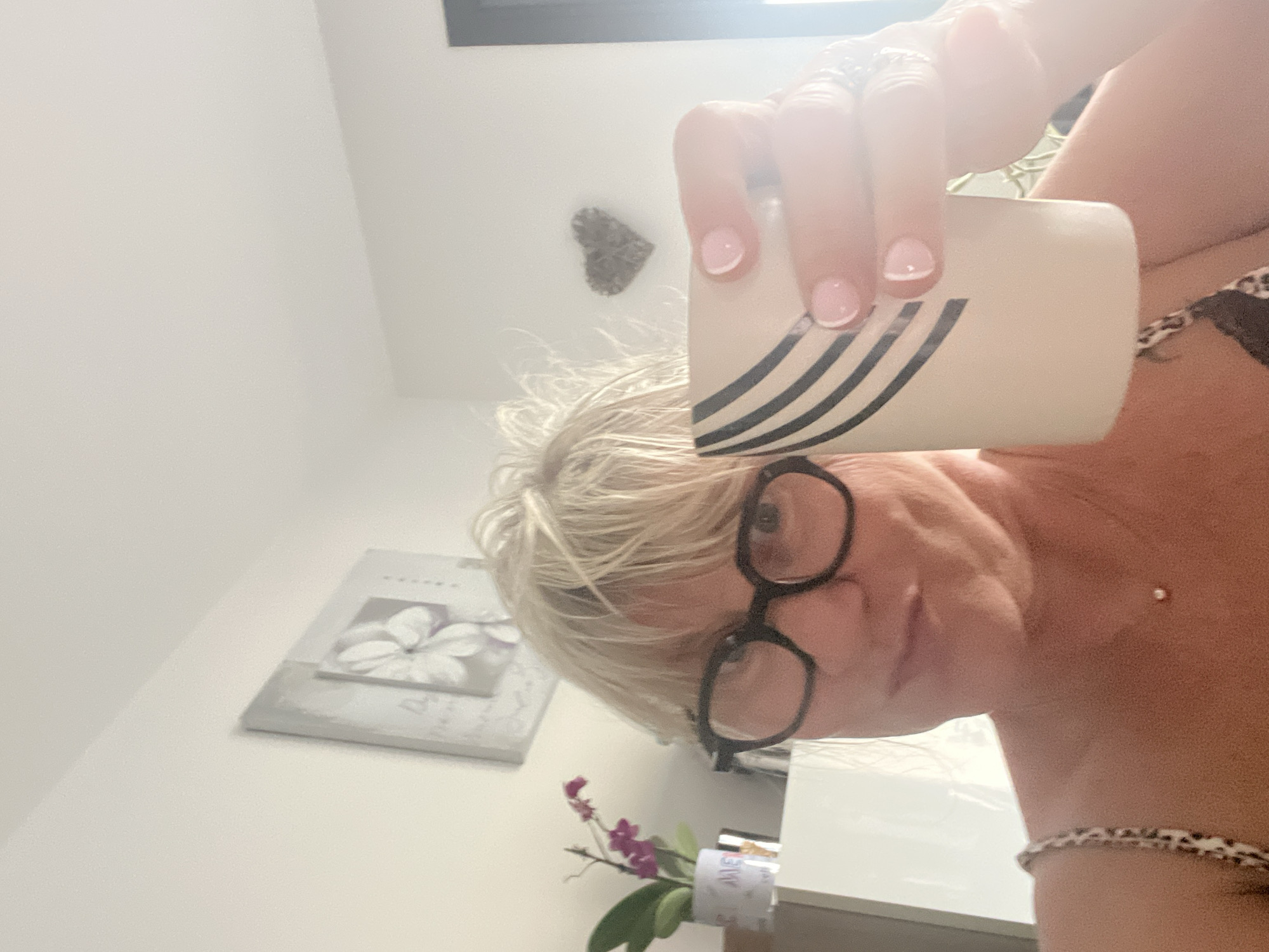The image shows a woman with blonde hair and glasses holding a cup. She is wearing a leopard-print top and a necklace with a small pendant. In the background, there is a picture of a flower on the wall and a decorative heart. There is also a potted orchid. The setting appears to be indoors, possibly a home, and the lighting suggests it is daytime. The location in Chamboeuf, France is not discernible from the image.