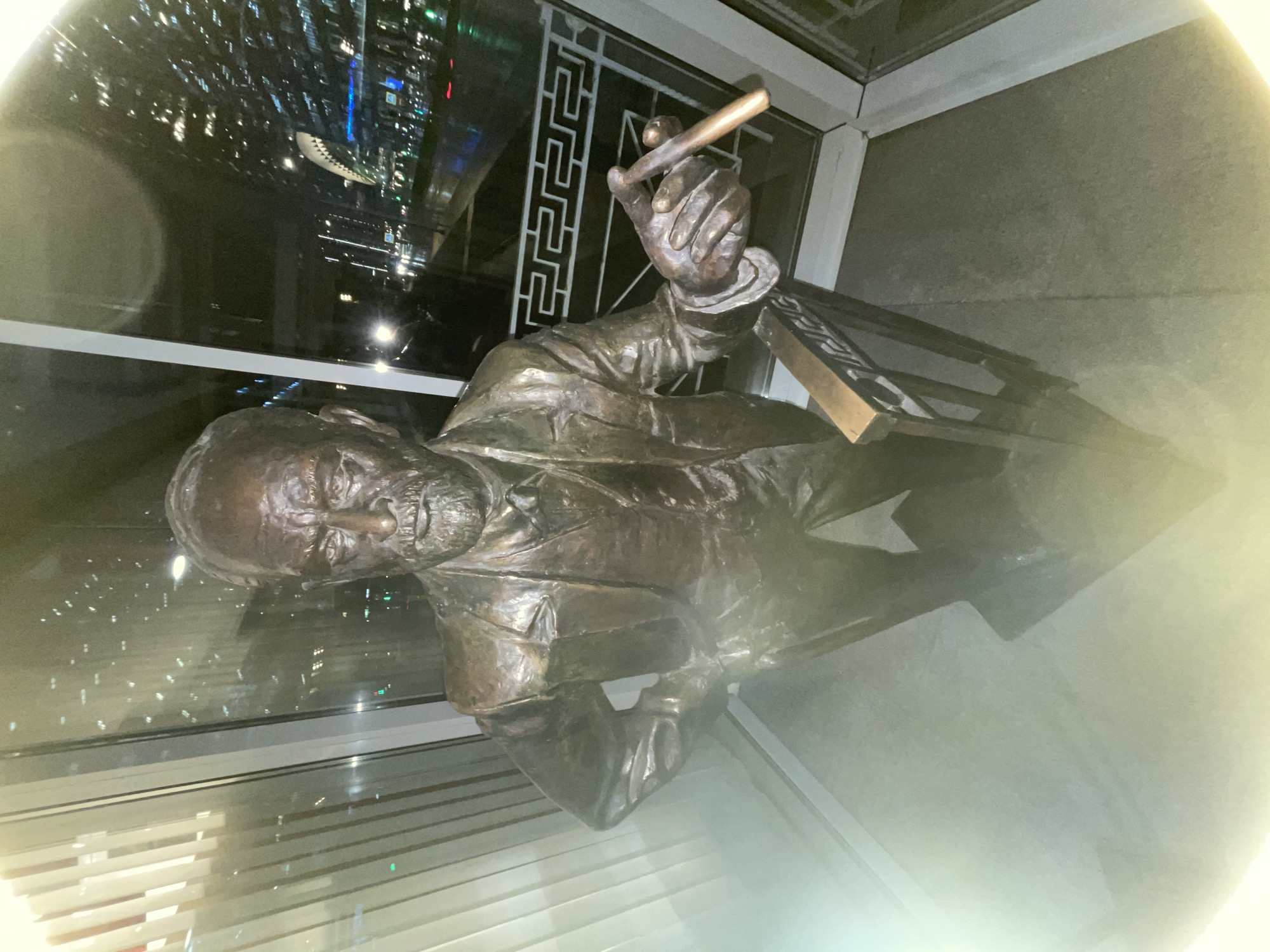 The image captures a bronze statue of Winston Churchill, cigar in hand, positioned indoors, seemingly within a building in Moscow, Russia. Reflected in the glass behind the statue are the lights of a modern cityscape at night. The statue is lit, with a halo-like effect surrounding it.
