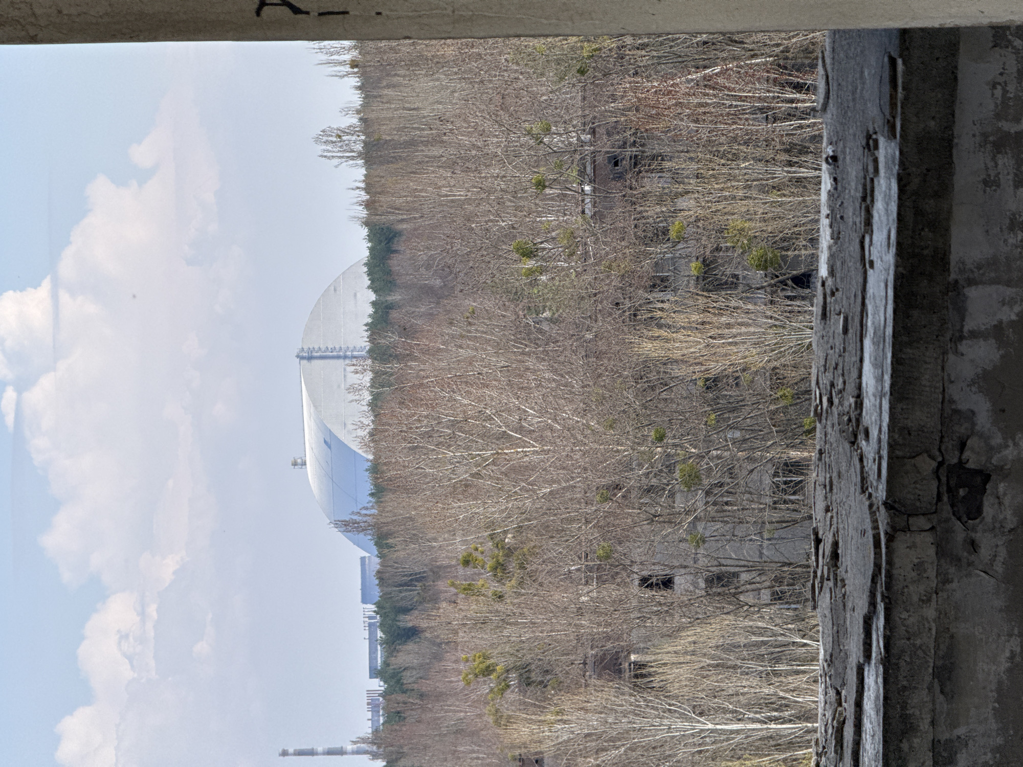 The image, taken from inside a dilapidated concrete structure, provides a partial view of the Chernobyl Nuclear Power Plant sarcophagus and the surrounding area in Prypiat, Ukraine. A dense, leafless forest fills the middle ground, partially obscuring abandoned buildings. The sarcophagus is visible to the left, appearing as a large, silver, semi-circular structure. The sky is light blue with white clouds. A partially framed view through a crumbling concrete structure is the closest foreground.