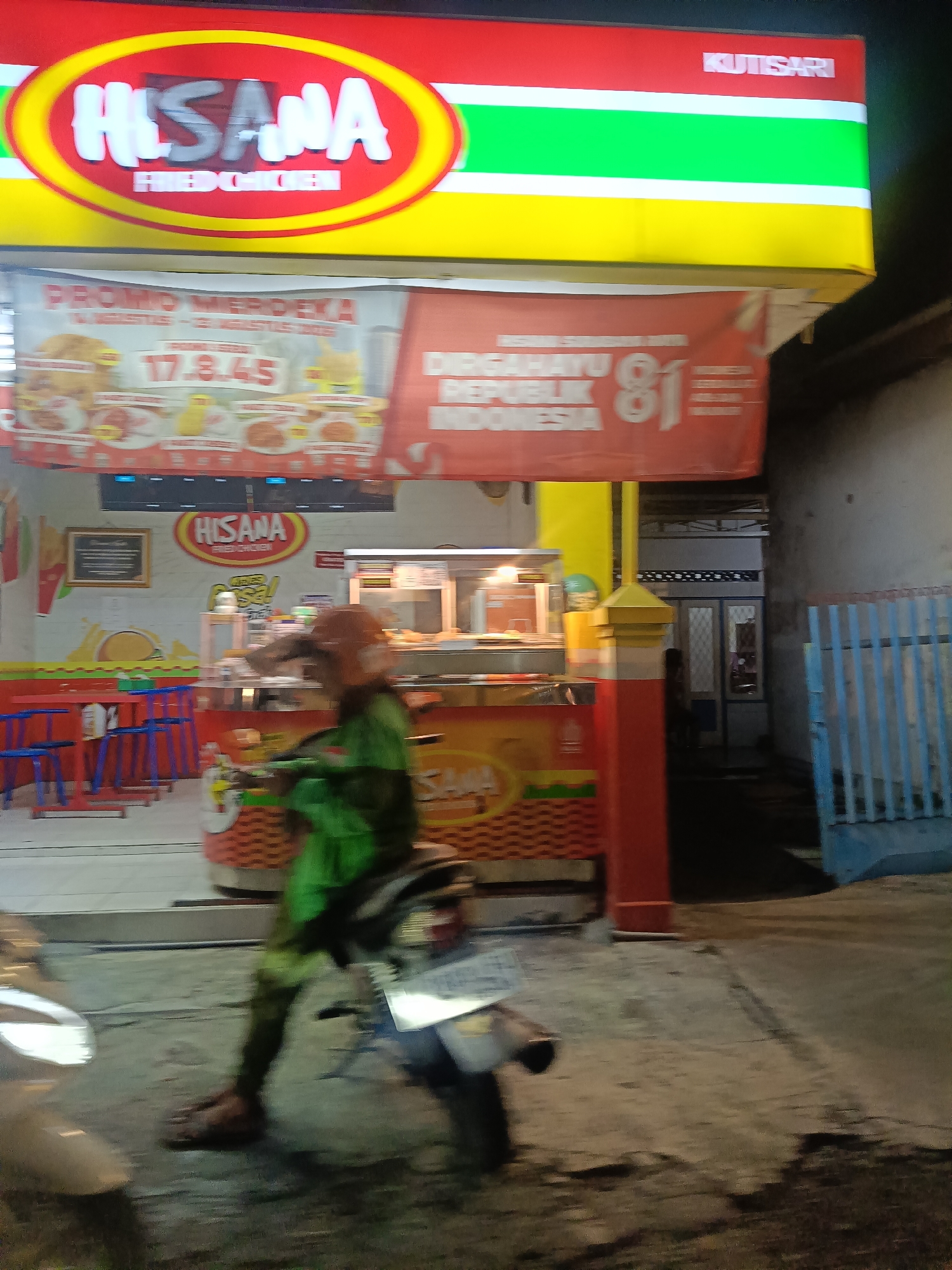 The image shows the exterior of a "HISANA FRIED CHICKEN" establishment in Surabaya, Indonesia, at night. A red, yellow, and green striped awning with the "HISANA" logo prominently displayed is visible. Below the awning, a banner proclaims "PROMO MERDEKA" and "DIRGAHAYU REPUBLIK INDONESIA 81", indicating a national independence day promotion. The establishment has red tables and blue chairs set up outside. A person wearing a green uniform and helmet is blurred due to motion, riding a motorcycle. The ground is paved with asphalt, and a blue metal fence is to the right. The lighting suggests it is after dark.