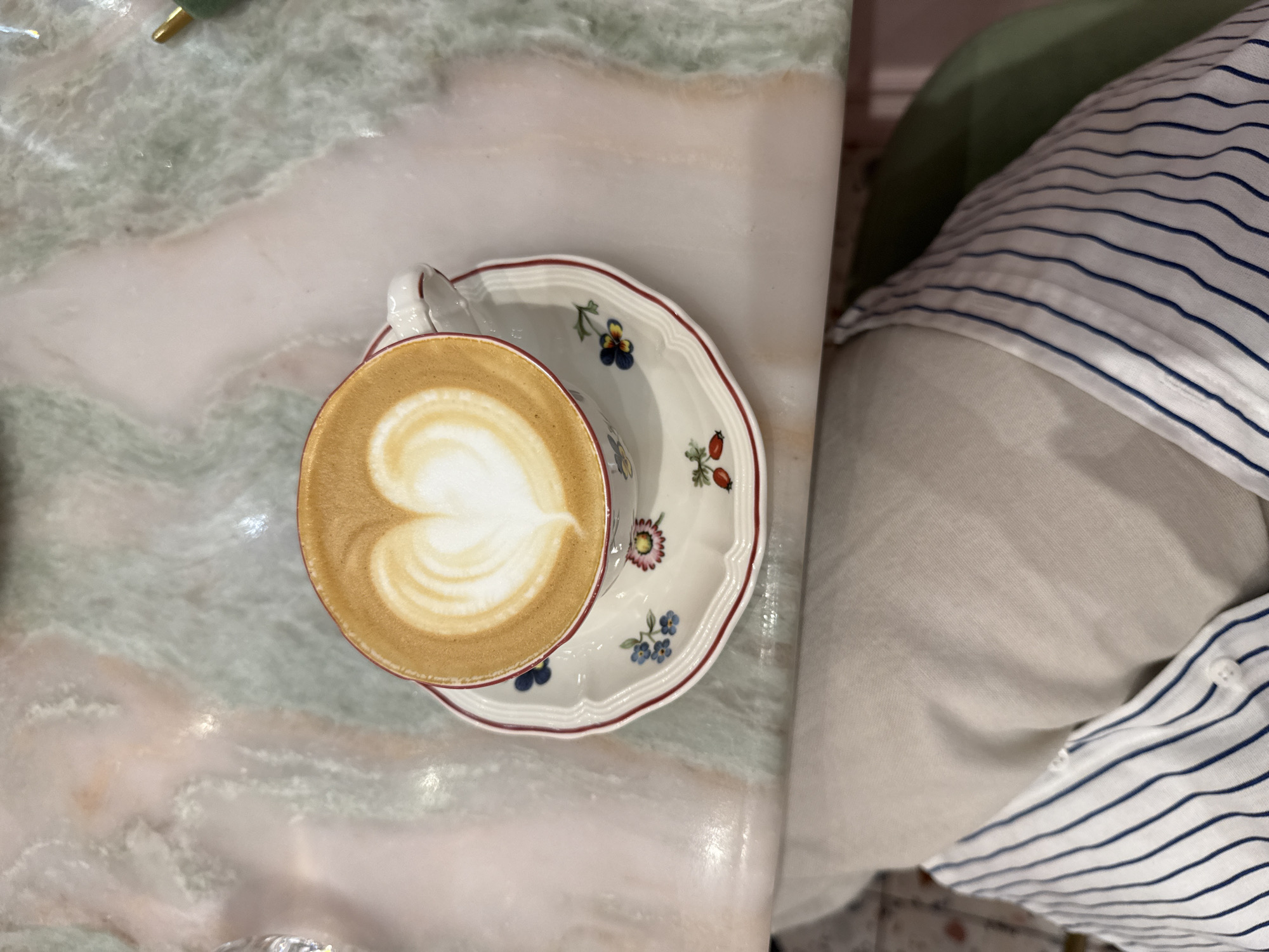A top-down shot showcases a cup of coffee with latte art in the shape of a heart, placed on a saucer with floral and berry designs. The coffee cup and saucer sit on a marbled surface. Below the table, a person's lap is visible, clad in a light-colored shirt with thin blue stripes and a button closure. A small section of a green object is visible to the right of the coffee cup. The scene is set indoors, with no specific location in the city of Unknown or Unknown being discernible.