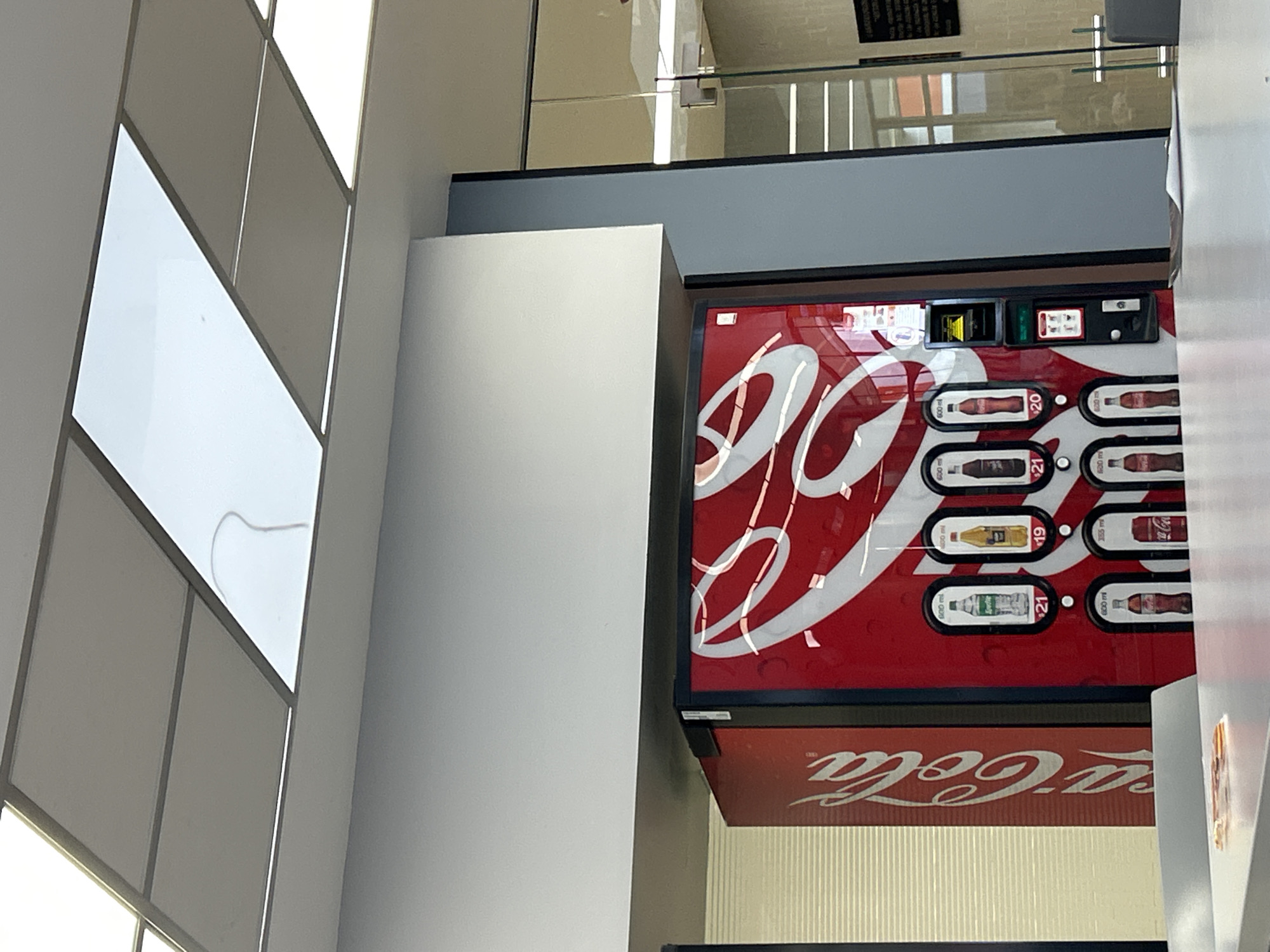 The image, rotated 90 degrees clockwise, captures an indoor commercial or public space featuring a prominent red Coca-Cola vending machine. The vending machine dominates the right side of the frame, showcasing rows of beverages. Visible products include various Coca-Cola bottles, a yellow-labeled bottle, and a Sprite bottle, each priced at "$20", "$19", or "$21" with "600 ml" indicated for most. An input slot for currency and a card reader are visible at the top of the machine.
To the left of the vending machine is a tall, white rectangular column or wall. Above and behind the vending machine, a glass railing suggests a mezzanine or upper floor, with reflections visible on its surface. Further left, a paneled ceiling with integrated rectangular light fixtures runs diagonally across the upper left portion of the image. Below the main vending machine, another red surface with part of a "Coca-Cola" logo is visible, possibly indicating another vending unit or a sign.
The scene is static, with no people or activities taking place. The time of day is not discernible due to the indoor setting and artificial lighting. The overall environment appears clean and modern, typical of a well-maintained building interior such as a lobby, cafeteria, or hallway.