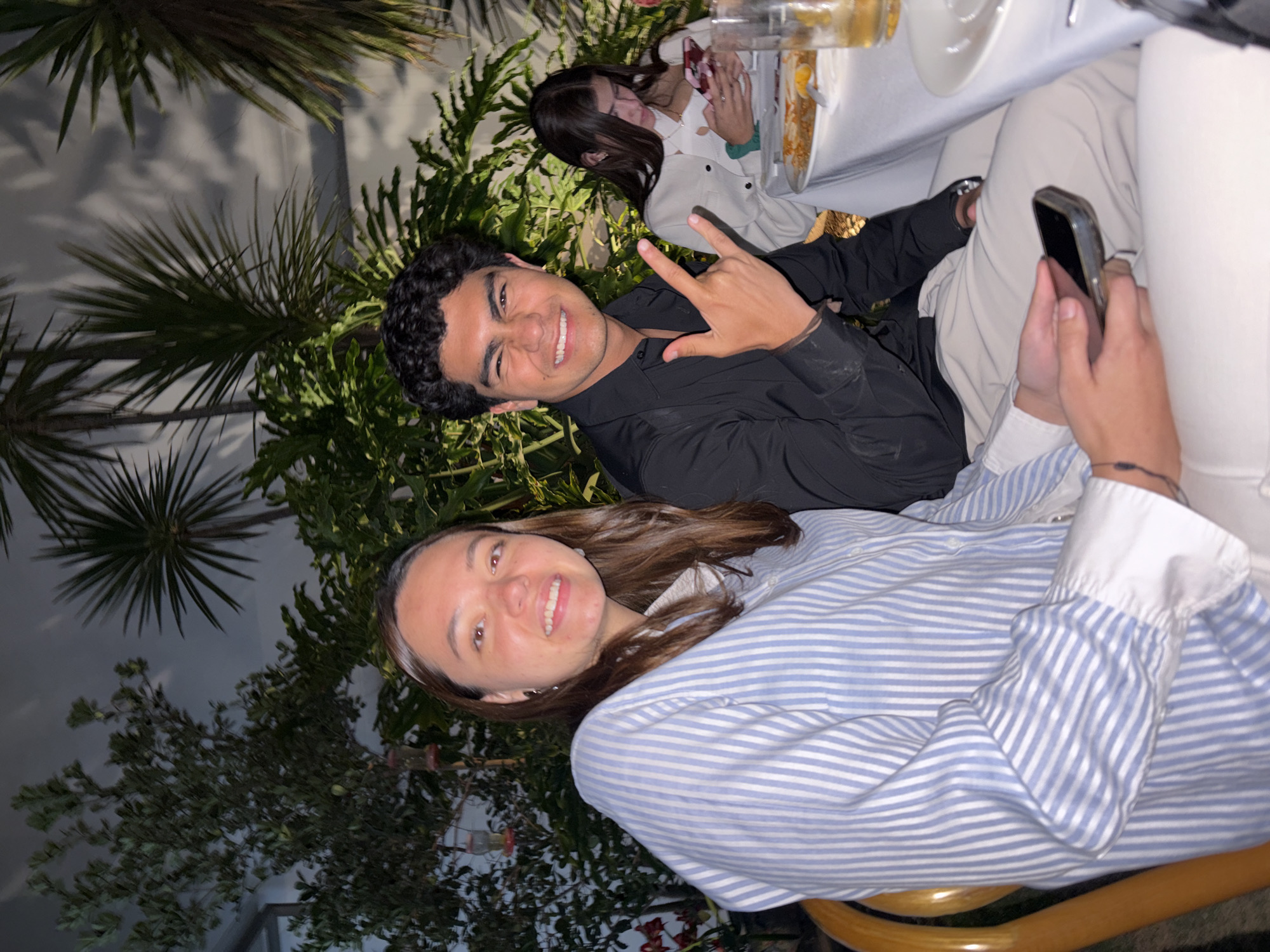 The scene depicts three individuals seated at an outdoor or semi-outdoor dining area at night in Puebla, Mexico.
The foreground features a young adult woman on the left, with long brown hair, wearing a light blue and white vertical striped button-up shirt. She smiles directly at the camera, holding a dark-cased smartphone with both hands. To her right sits a young adult man with dark, curly hair, dressed in a black long-sleeve button-up shirt and light-colored trousers. He also smiles directly at the camera, forming a "rock on" gesture with his raised right hand.
In the right background, a third young adult woman, with long dark hair and wearing a light-colored top, is visible from the chest up. She is seated, focused downwards on a red-cased smartphone held in her hands.
A table covered with a white tablecloth is visible in the right midground, holding a plate containing food with orange and yellow elements, and a clear beverage glass. The background is dominated by dense, illuminated green foliage, including palm-like and broad-leafed plants, extending up to a dark, textured overhead surface. Shadows from the vegetation are cast onto this surface. A small, red and clear plastic object, resembling a bird feeder, hangs within the foliage in the upper left portion of the image.