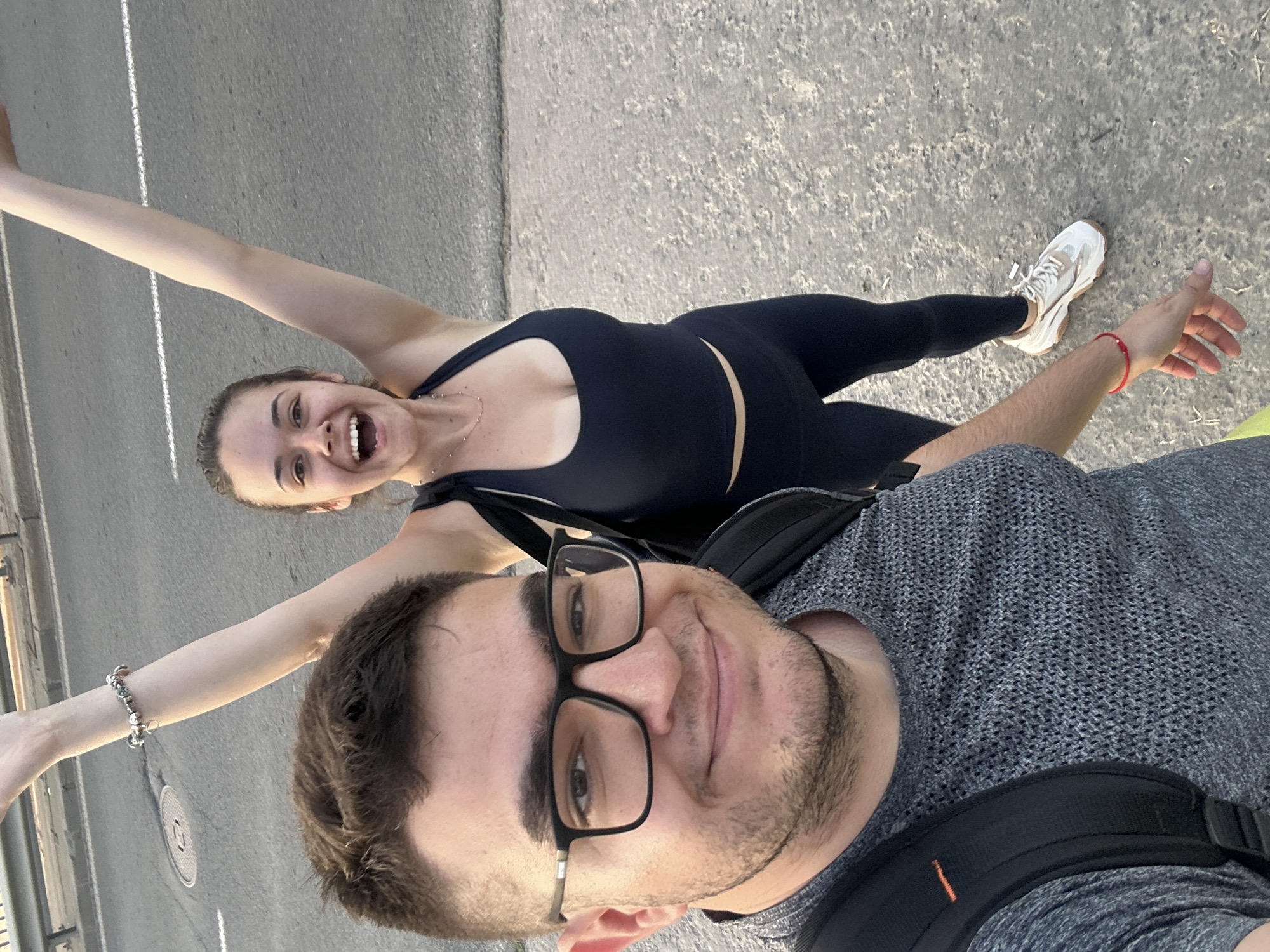 A medium shot shows a couple taking a selfie on a city street in Sofia, Bulgaria. The image is rotated counter-clockwise, placing the woman at the top and the man at the bottom.
The woman, fair-skinned with brown hair pulled back, is wearing a black athletic top and leggings. She is smiling broadly with her arms outstretched. She is wearing white sneakers and a silver bracelet on her left wrist.
The man, with fair skin, dark hair, a beard, and glasses, is smiling. He is wearing a gray knit shirt and a backpack with black straps.
The background shows a gray asphalt street. A manhole cover and white lane markings are visible. There is a partial view of a concrete barrier on the left side.