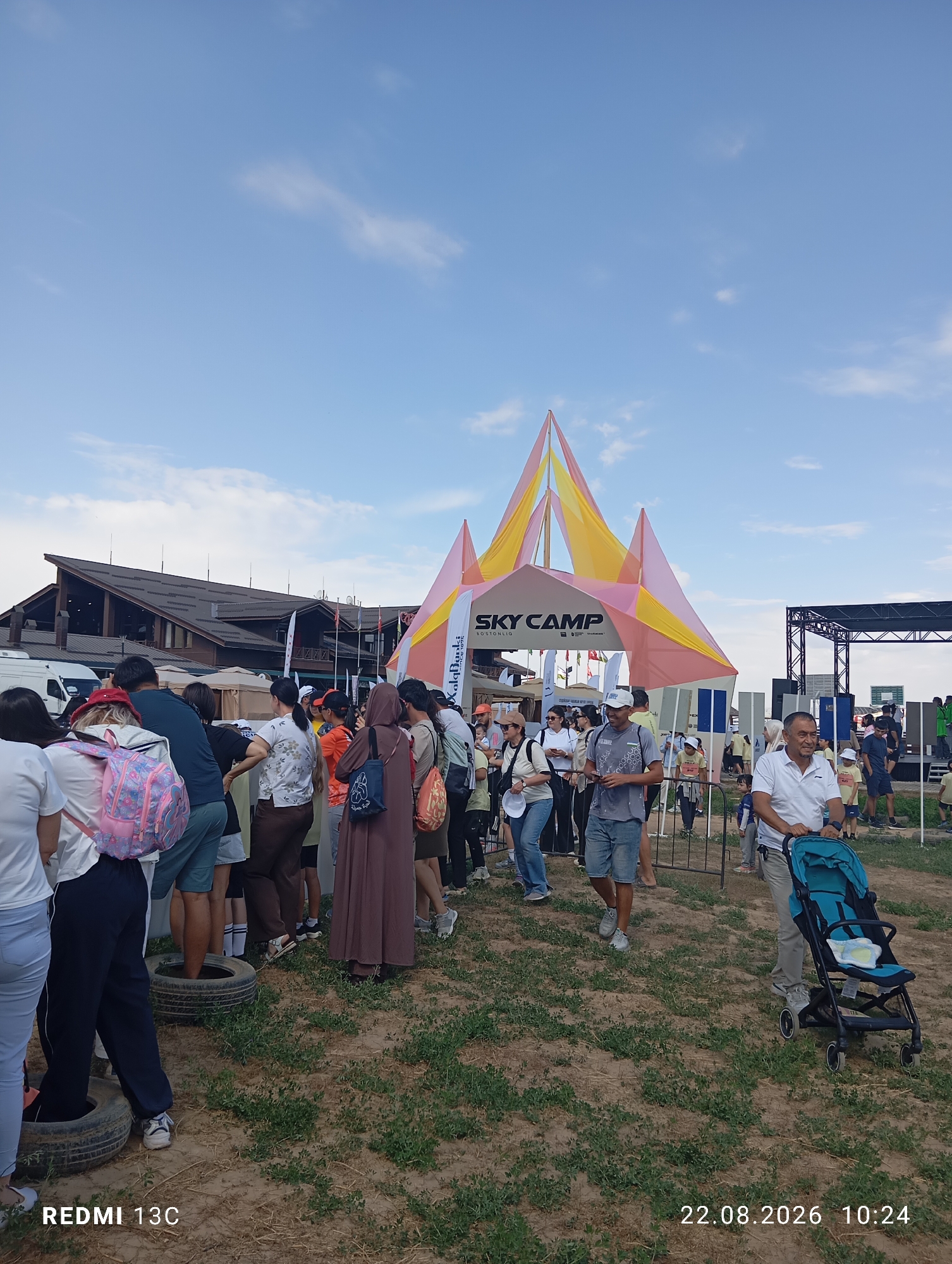 The image depicts a busy outdoor event at the Amirsay ski resort in Uzbekistan. In the foreground, a group of people are gathered, some waiting in line. The "Sky Camp" structure, a brightly colored tent with yellow and pink fabric, serves as a central point. Behind it, a large building, likely part of the ski resort facilities, is visible. Various flags are displayed, suggesting an international or organized event. To the right, a stage structure is set up, and more people are observed in the background, indicating a lively atmosphere. A stroller is being pushed by a man on the right side of the frame. The ground is covered with dry grass and some dirt. The sky is blue with scattered clouds. The date and time "22.08.2026 10:24" and "REDMI 13C" are watermarked on the bottom of the image.