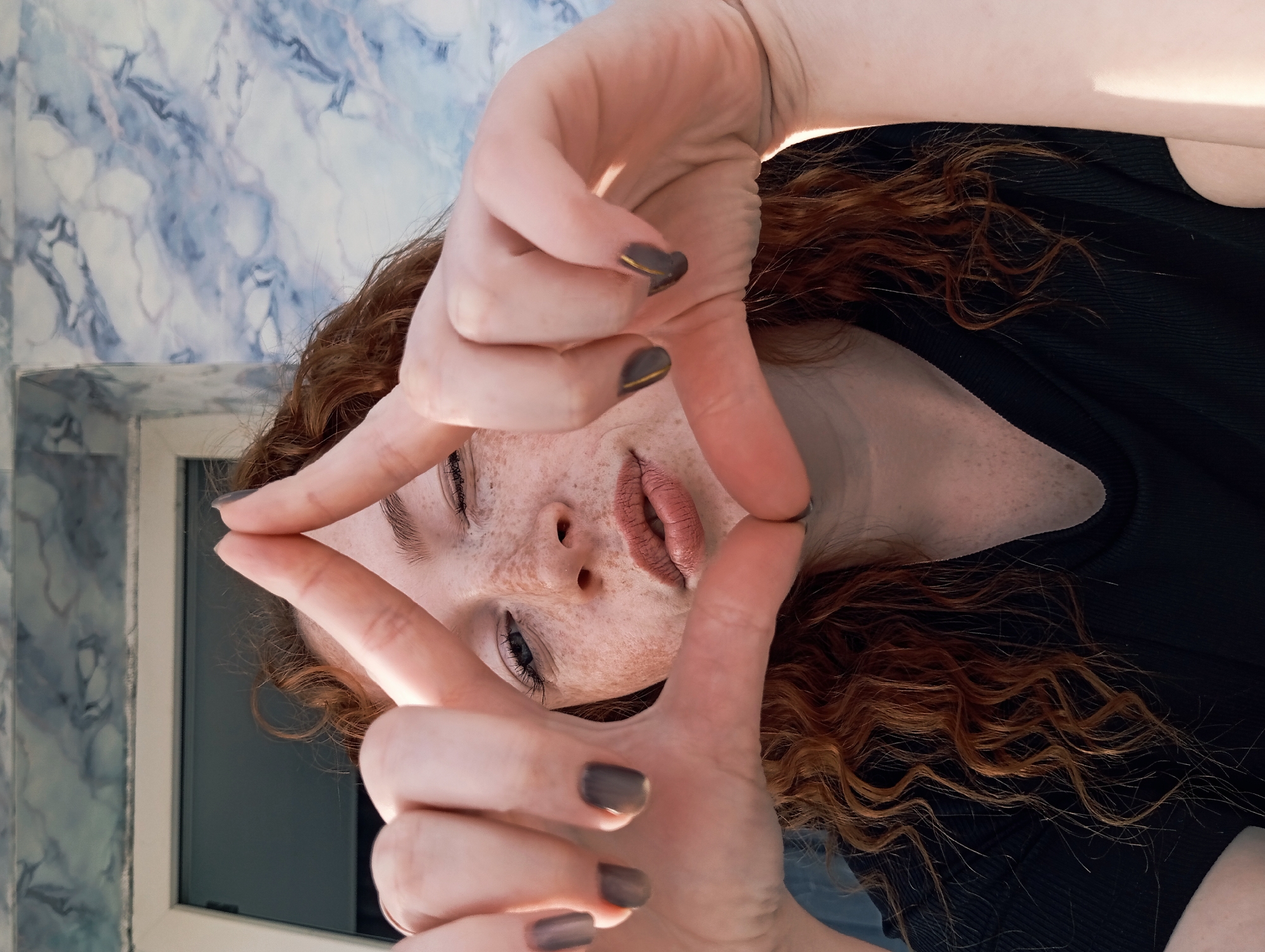 A close-up, portrait-style image features a young individual with fair skin, numerous freckles across their nose and cheeks, and long, wavy red hair. The person is framing their face with both hands, creating a rectangular shape. Their fingernails are painted a dark, possibly charcoal grey or brown, each adorned with a thin gold horizontal stripe near the tip.
The individual's eyes are open, looking directly at the viewer, and their lips are slightly parted. They are wearing a dark, presumably black, ribbed garment. The background consists of a light blue and white marbled wall, with a segment of a window frame visible in the lower left, looking out onto a dark area. The lighting suggests an indoor setting, with some highlights on the hair and skin. No text is visible in the image. This scene is located in Yurgamysh, Russia.
