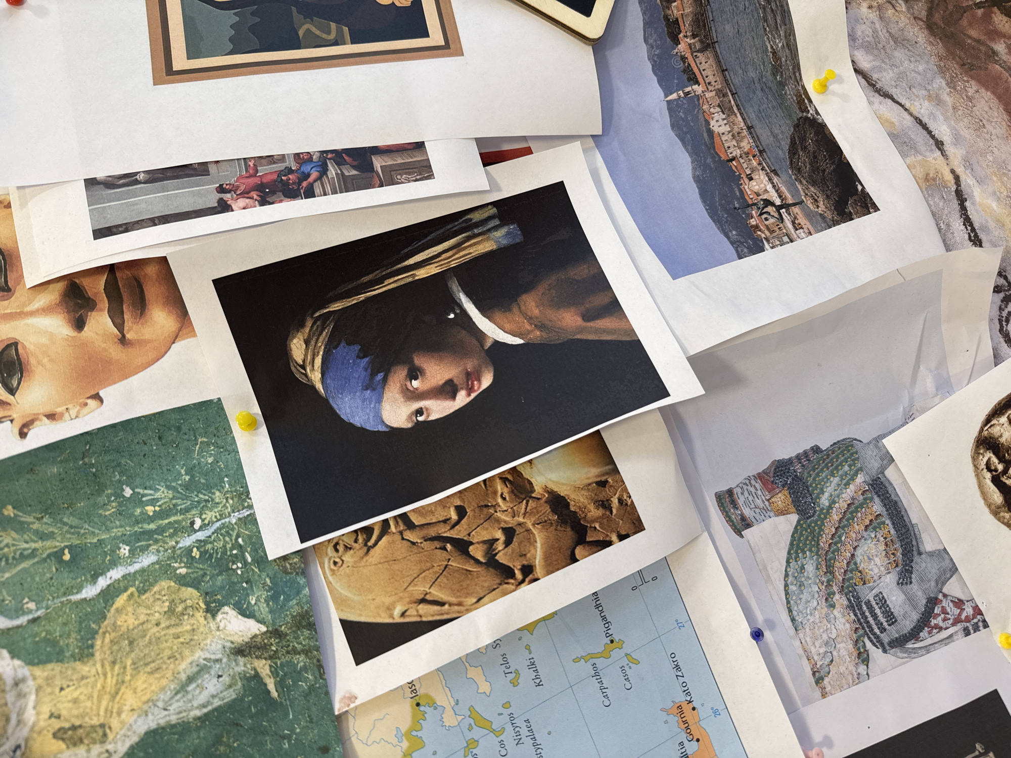 The image shows a collection of printed images pinned to a surface. A print of Johannes Vermeer's "Girl with a Pearl Earring" is prominently displayed. Other images include what appear to be artistic renditions or photographs of artwork, architecture, and maps. There are visible pushpins in yellow, blue, and red colors used to secure the images. The map appears to depict the Eastern Mediterranean, including islands like Cos and Carpathos. There is no visible connection to Budva, Montenegro.