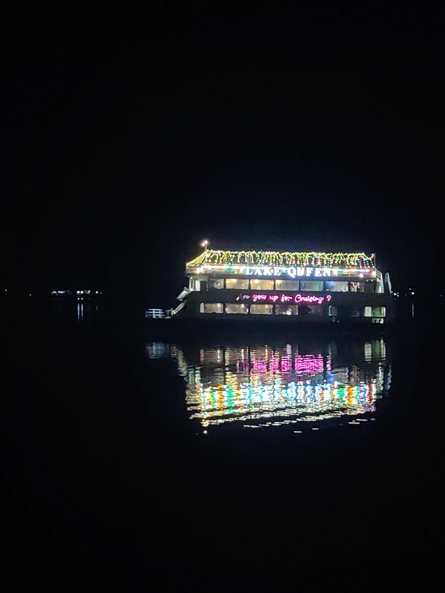 The image shows a boat named "LAKE QUEEN" illuminated with colorful lights on a body of water at night. The boat is decorated with strings of lights along its roof and sides. A neon sign on the side of the boat reads "Are you up for Cruising?". The lights from the boat are reflected in the dark water. In the distance, on the left side of the image, there are other faint lights on the water, possibly from other boats or structures. The overall scene appears festive and serene. This boat, "LAKE QUEEN," is likely operating on Ramgarh Tal in Gorakhpur, India, a popular destination for boating and recreation.
