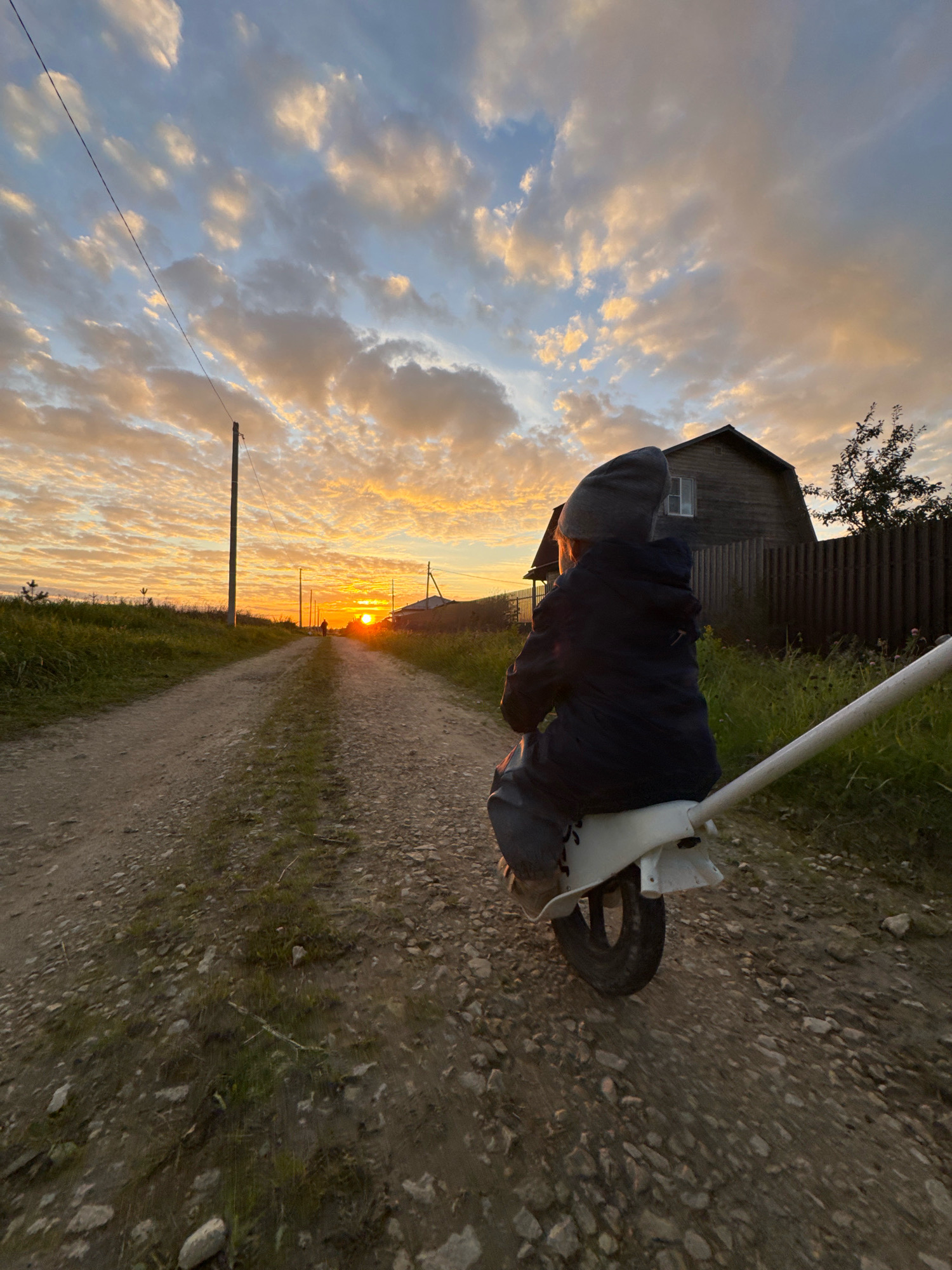 An over-the-shoulder shot shows a young child, wearing a dark jacket and a grey beanie, sitting on a white unicycle. The child is facing away from the camera, towards a sunset in Kempelevo, Russia. The unicycle is positioned on a dirt road, flanked by tall grass and small trees. In the background, a wooden fence and a rustic house are visible, partially obscured by the setting sun. The sky is filled with dramatic clouds, illuminated by the warm hues of the sunset. A power line stretches across the frame from the left, supported by a wooden pole.