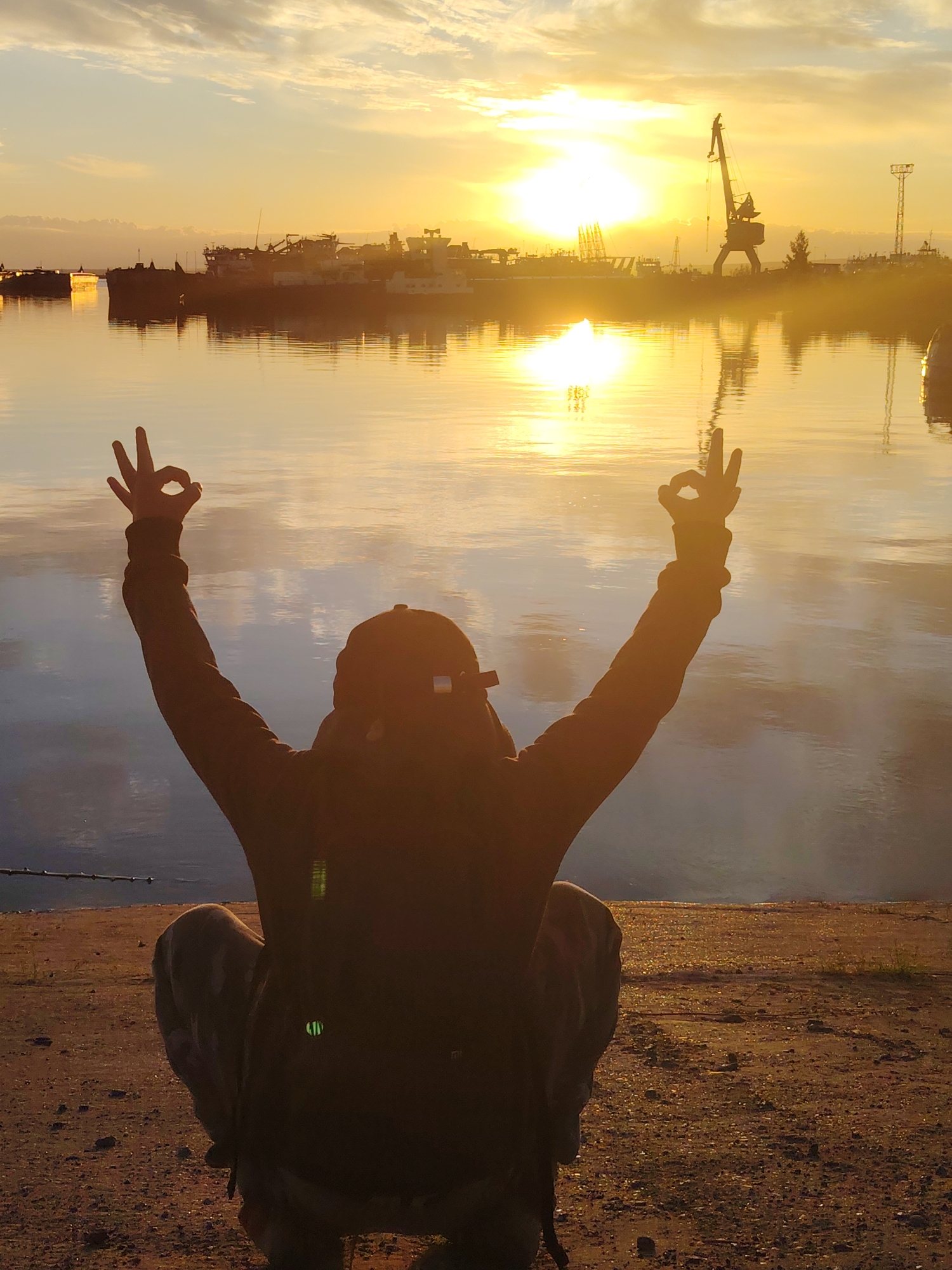 A person crouches on a gravelly shore, facing away from the camera, with arms raised and hands forming the "okay" gesture. The scene is illuminated by the golden light of a sunrise or sunset over a body of water, likely the Kama River in Chaikovsky, Russia. The water reflects the sky and a silhouette of industrial structures, including a large crane, line the opposite bank. The person is wearing a hooded jacket and a baseball cap.